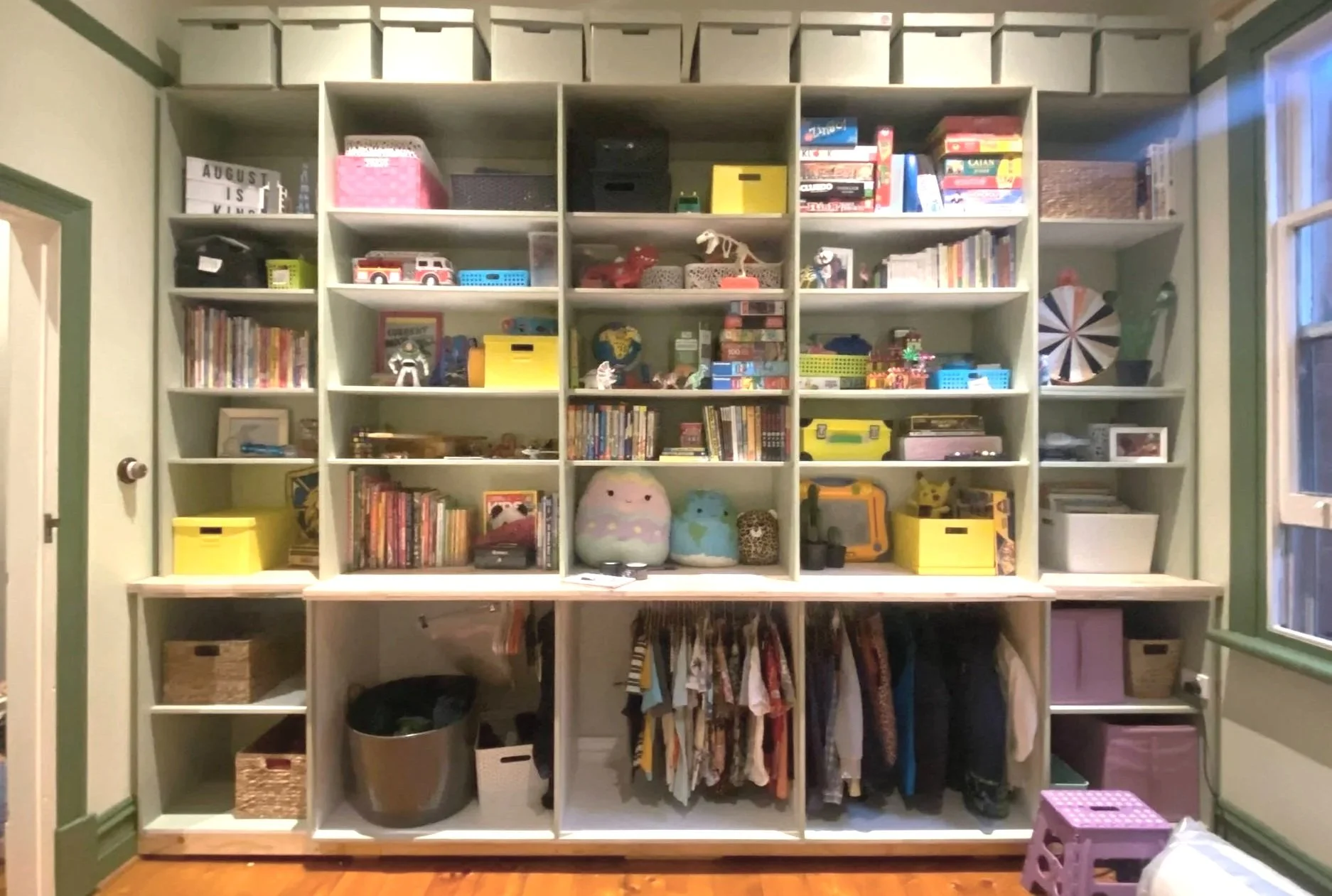 A white bookshelf filled with various toys, books, and storage containers, with a few items on top and a small pink stool in front.