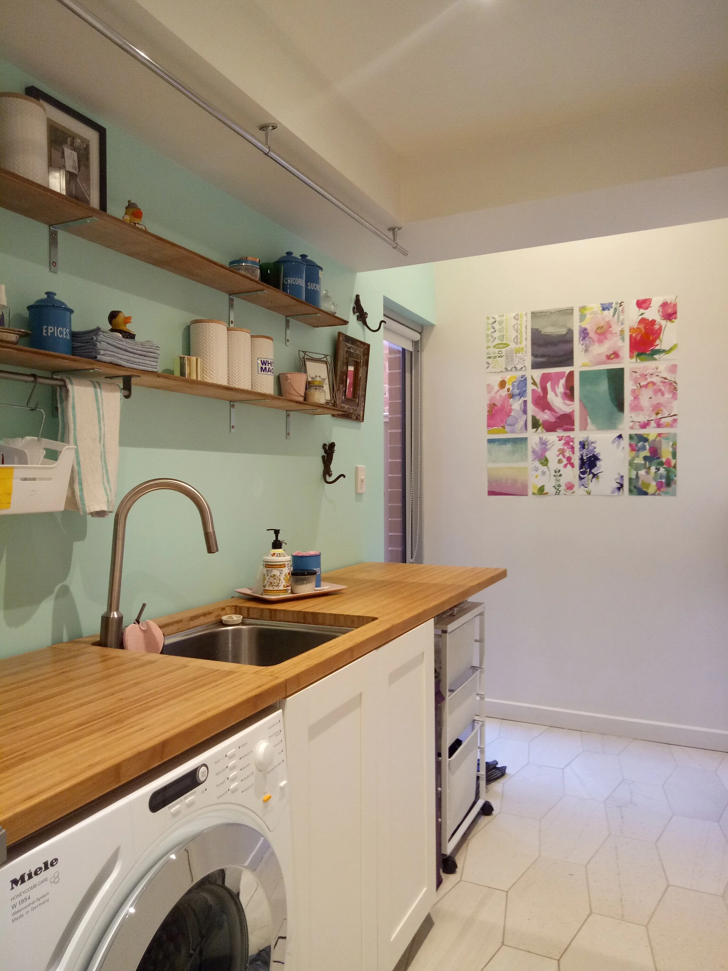 Kitchen with a laundry area, featuring a wooden countertop, a sink, shelves with containers and decorative items, and a wall with colorful floral artwork.
