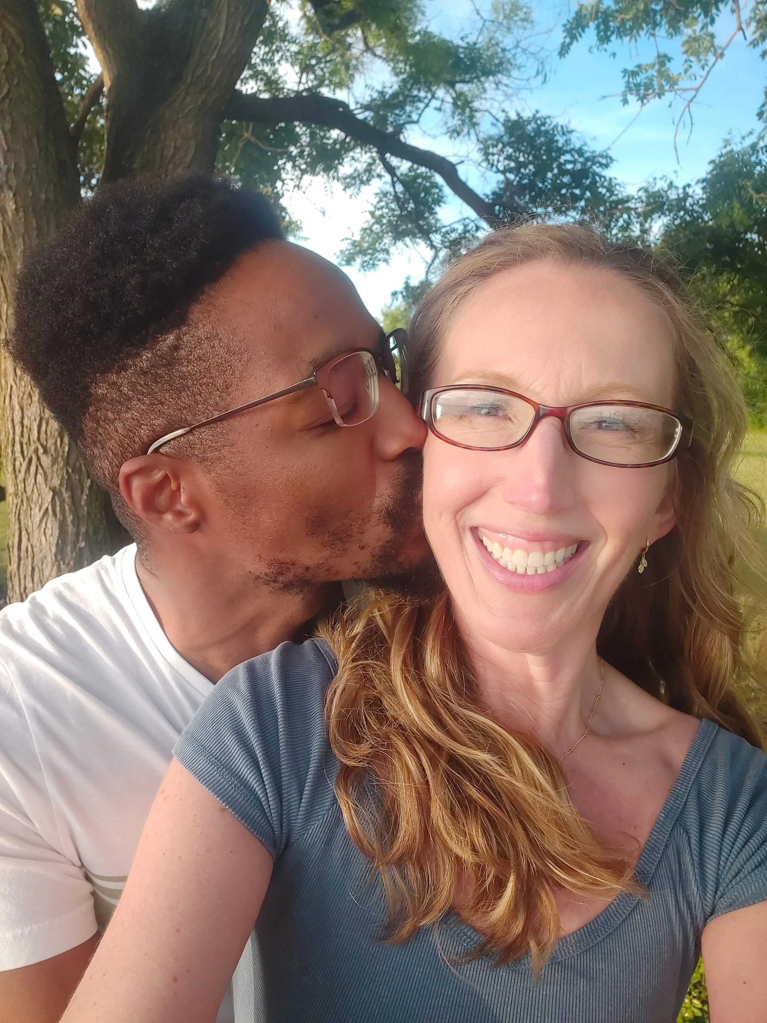 A smiling woman with glasses and long red hair taking a selfie outdoors, with a man kissing her cheek behind her, under a tree with green leaves and blue sky in the background.