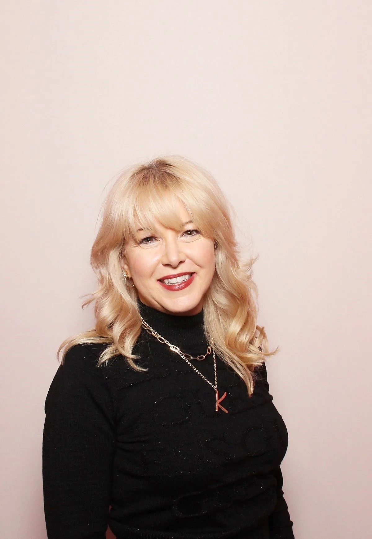 A woman with blonde, curly hair, smiling, wearing a black turtleneck sweater and layered necklaces, standing against a plain off-white background.