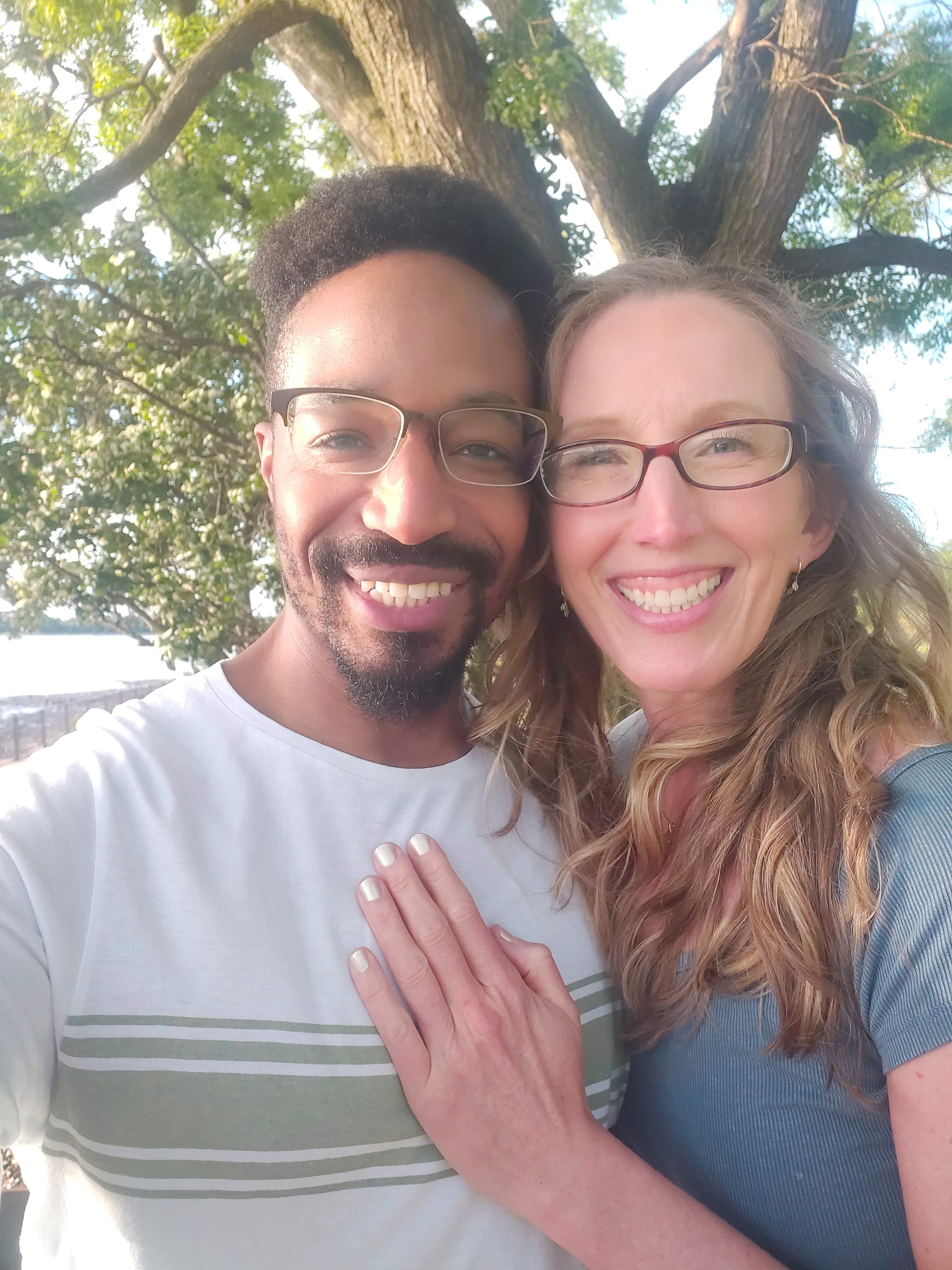 Close-up of a smiling young couple, a man with glasses and short dark hair, a woman with glasses and long wavy light brown hair, standing outdoors near a tree with green leaves, during daytime.