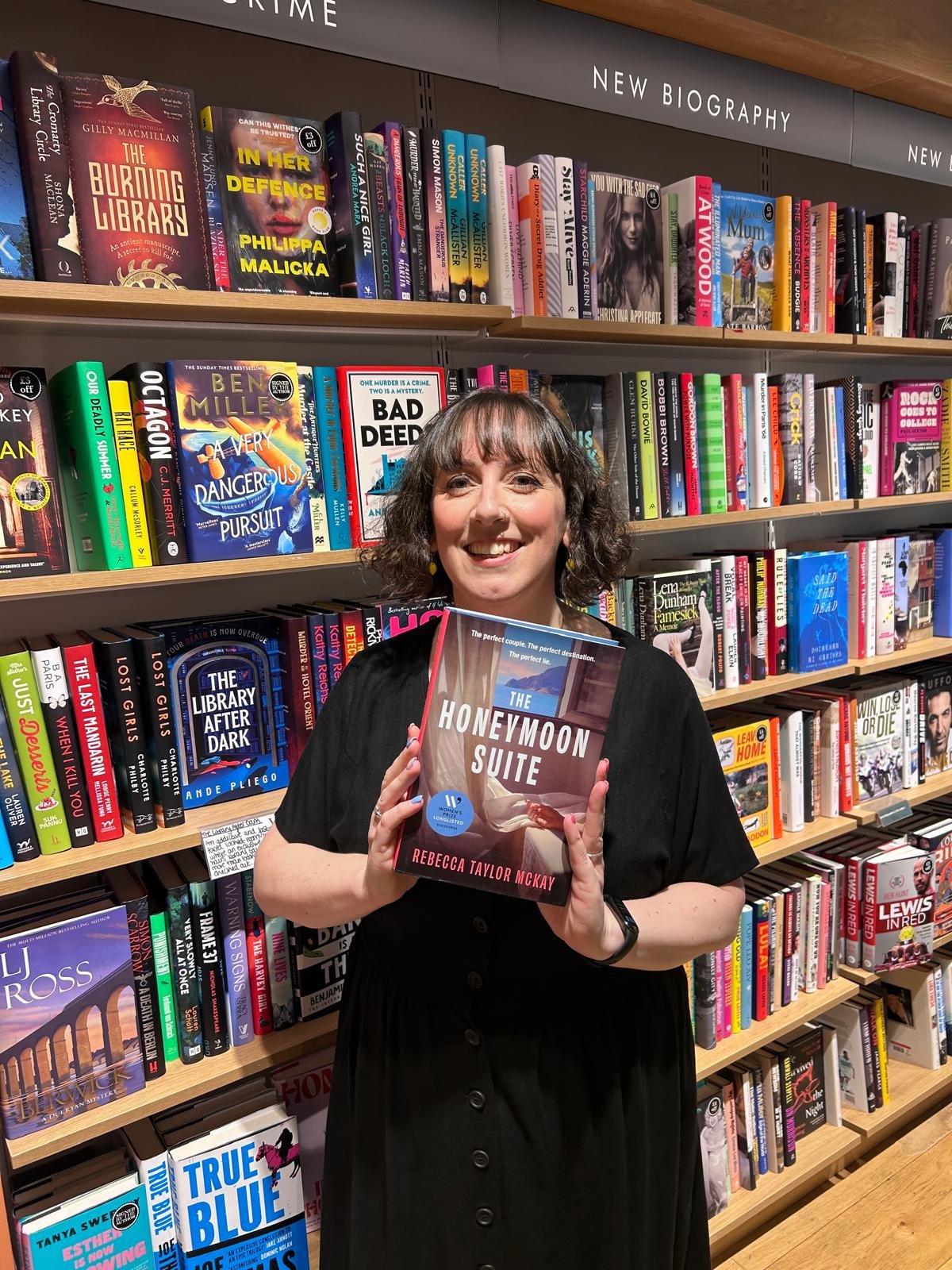 the image shows Rebecca Taylor McKay standing in front of bookshelves in a bookshop, holding a hardback copy of The Honeymoon Suite and smiling