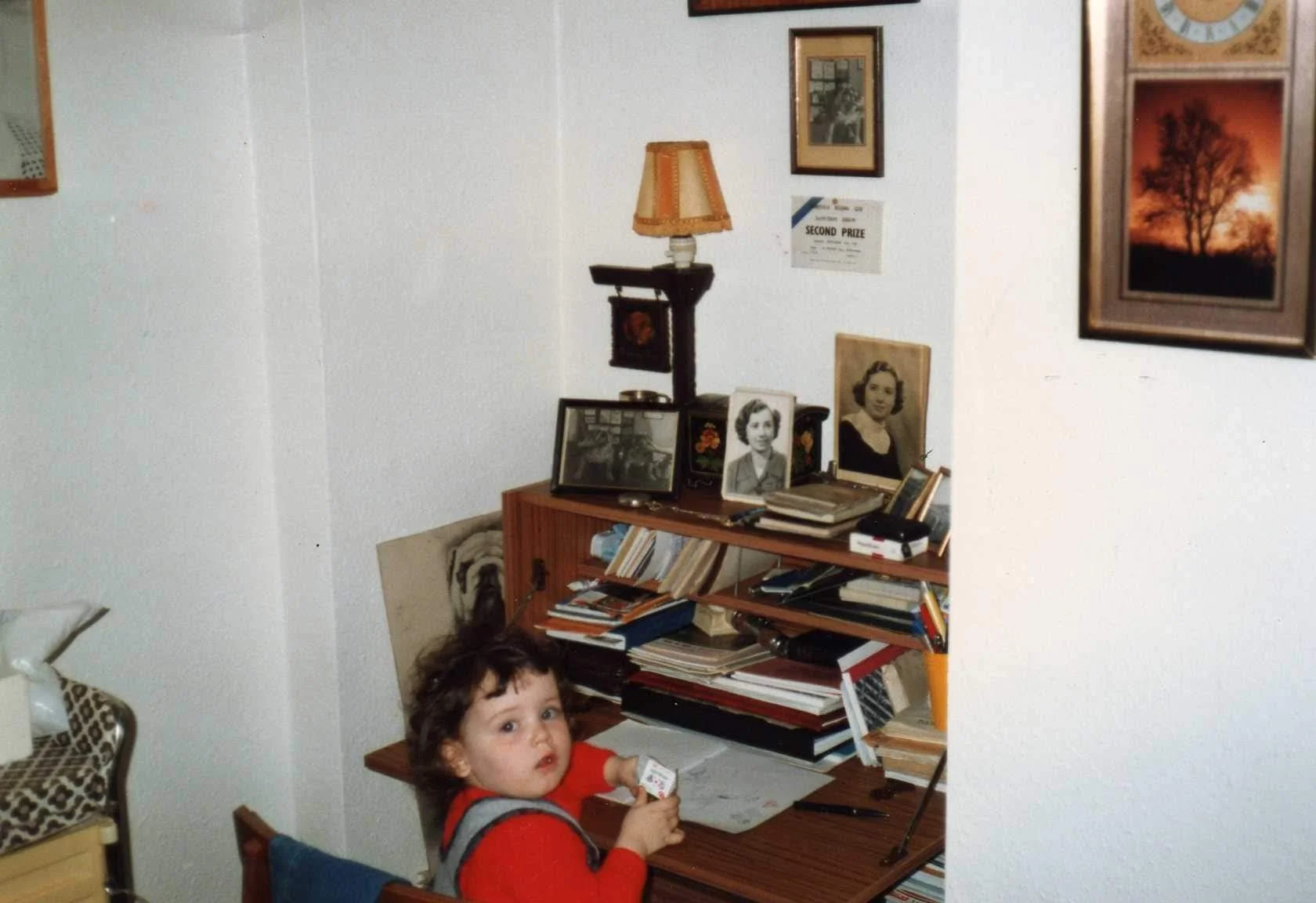 Image shows a baby with white skin and black hair wearing a red jumper and overalls, sitting up to a drop-leaf desk stacked with notebooks. Above the desk, a small certificate reads 'SECOND PRIZE'