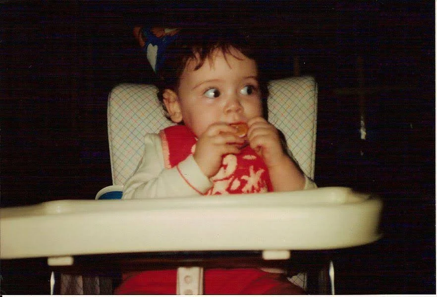 Image shows a baby with white skin and black hair and wide, dark eyes sitting in a highchair, wearing a cone-shaped party hat and sucking on an orange segment