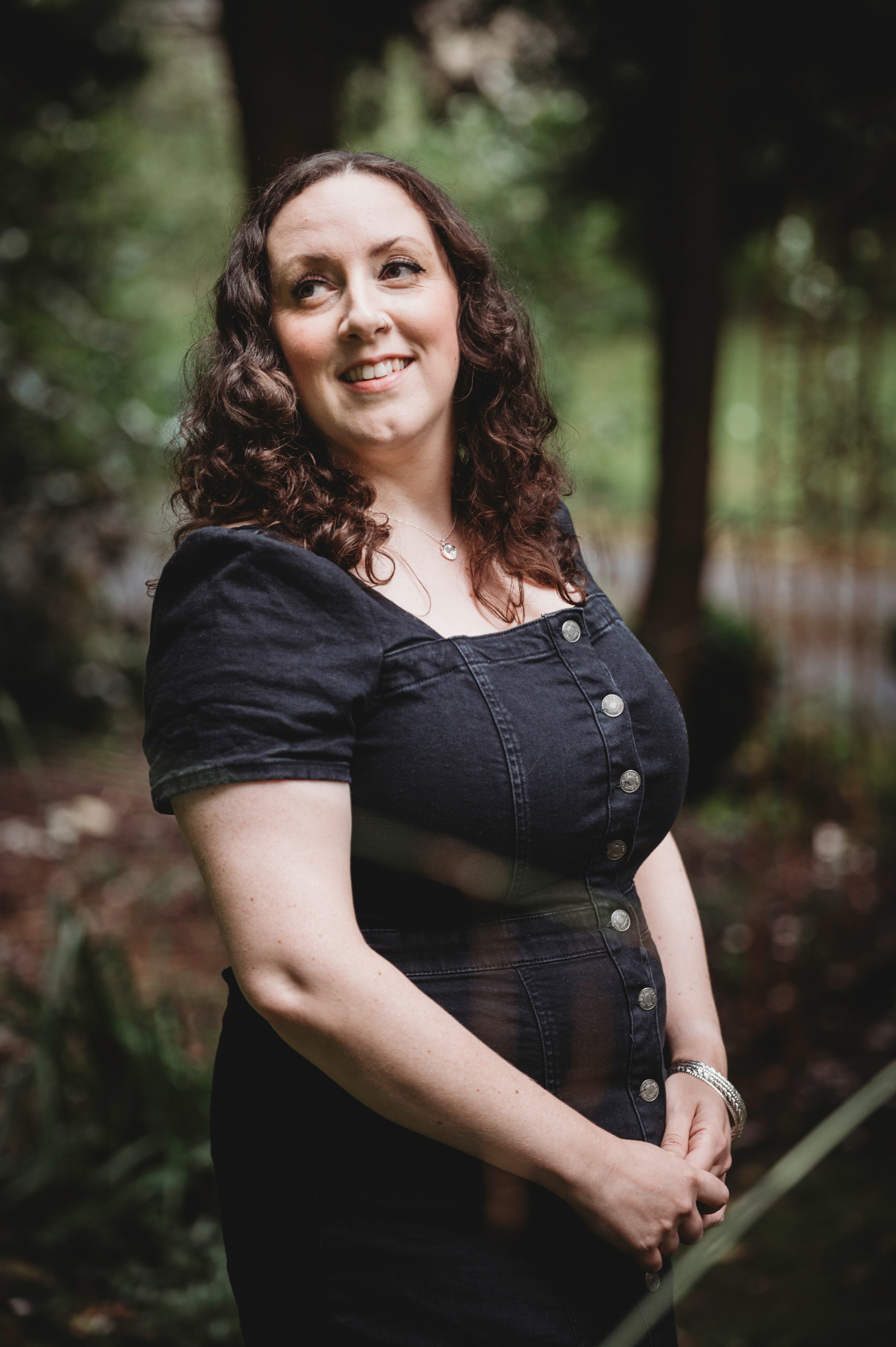 Image shows the author - a white woman in her 40s with dark curly hair, standing in shaded woodland, looking over one shoulder and smiling with her hands clasped in front of her