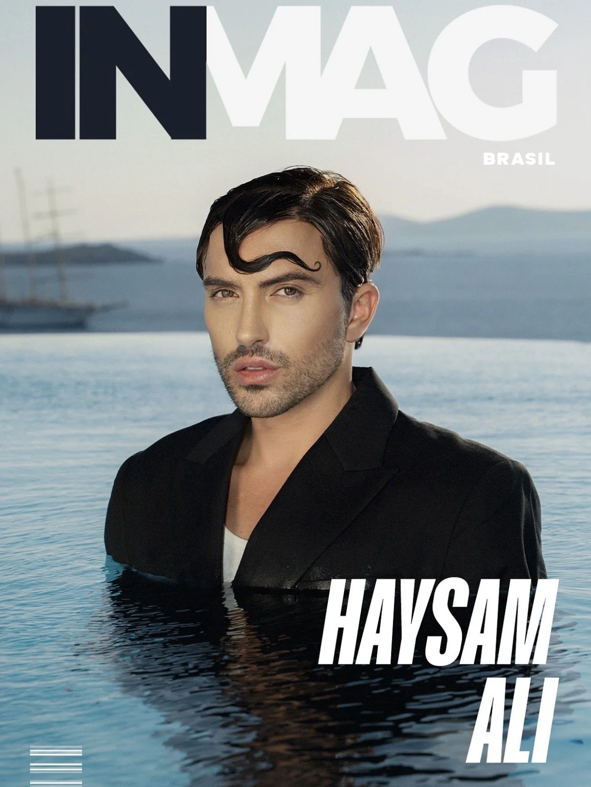 Magazine cover featuring a man with dark hair and a combed wave hairstyle, wearing a black blazer over a white shirt, standing in water with a scenic coastal background.