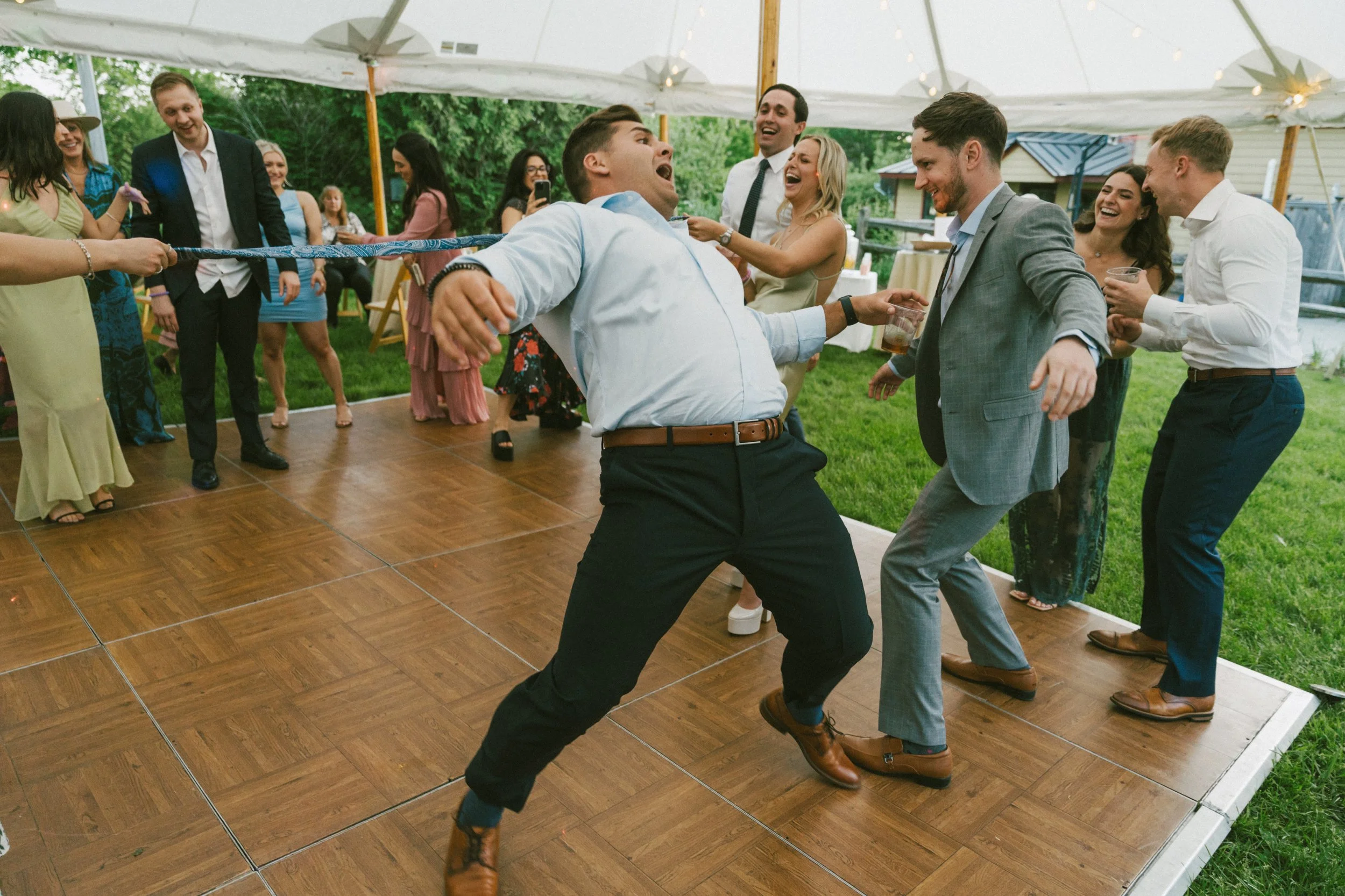 Wedding guests laughing and dancing under a tent during a lively outdoor reception, captured during a fun limbo game.