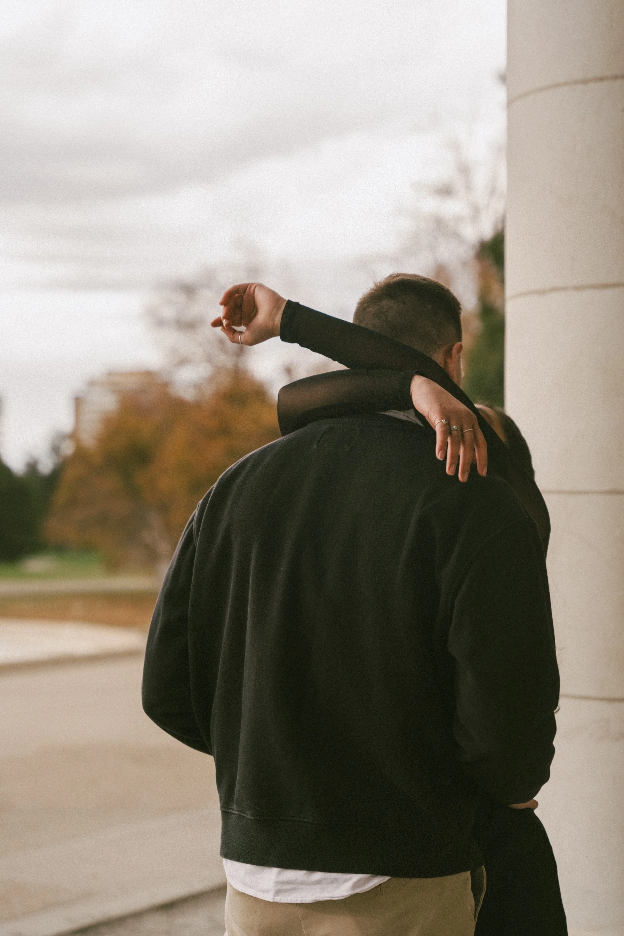 A couple embraces in front of cream colored pillars and orange foliage. The bride-to-be wraps her arms romantically around her groom-to-be's neck as they embrace for a romantic kiss.