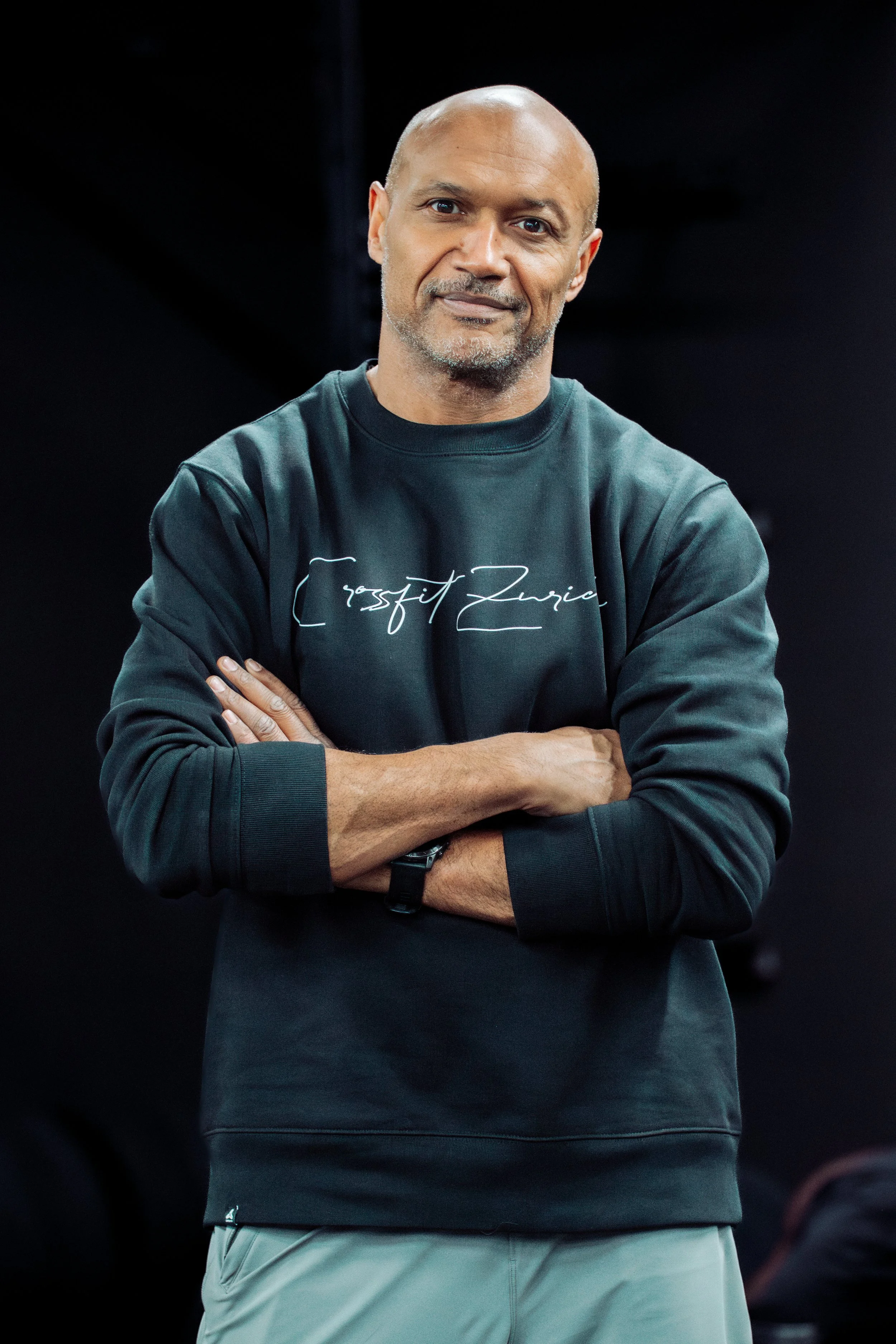 A man with a shaved head and goatee, wearing a black sweatshirt with white cursive writing, standing with his arms crossed in front of a dark background.