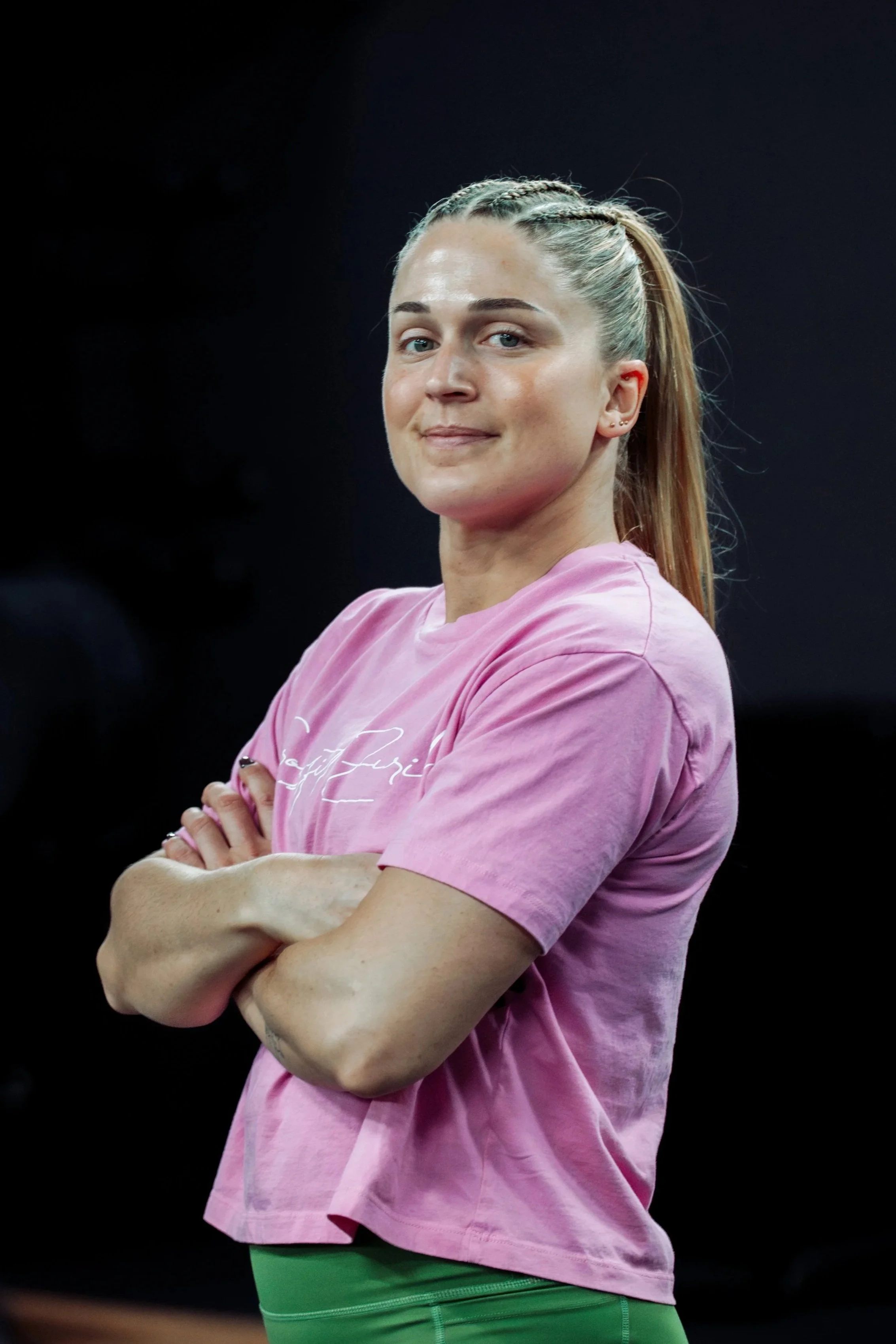 A woman with light skin, blonde hair styled in small braids, wearing a pink T-shirt and green leggings, standing with her arms crossed against a dark background.
