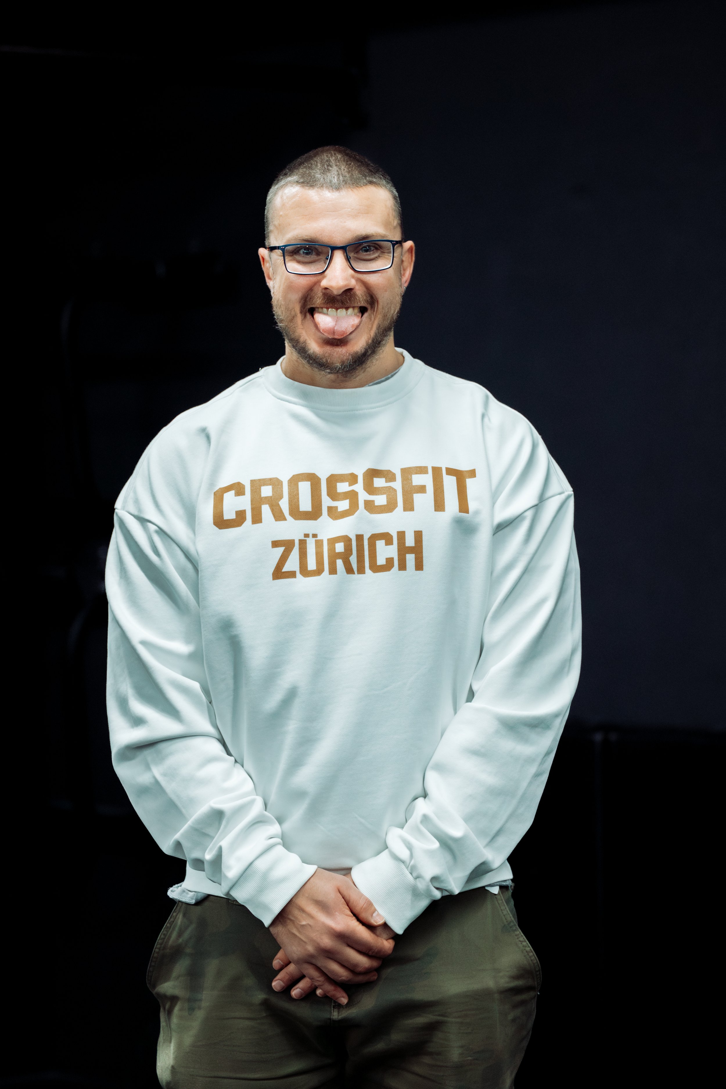 A smiling man with glasses and a short beard wearing a white sweatshirt with 'CROSSFIT ZÜRICH' printed on it, standing against a dark background.