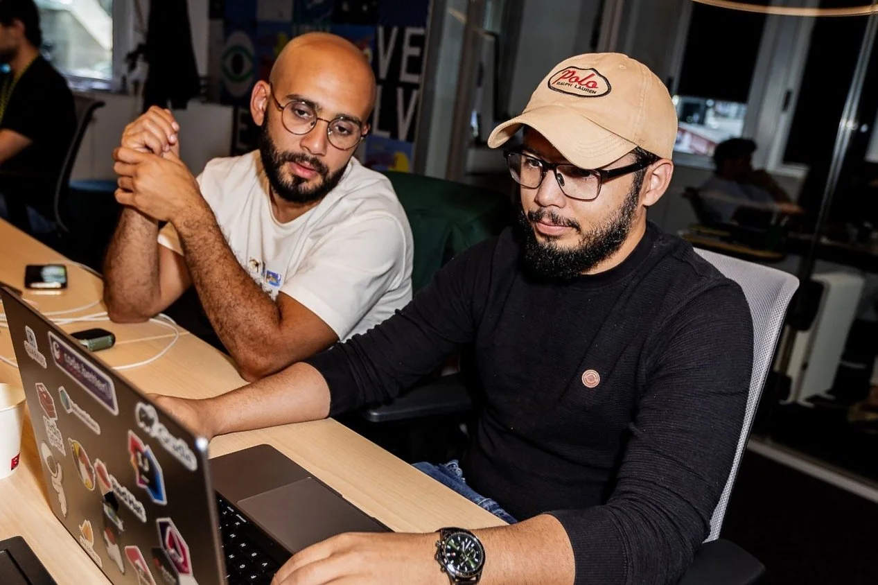 Two men collaborating at a desk with laptops, one wearing glasses and a black shirt, the other in a white shirt and round glasses, in an office setting.