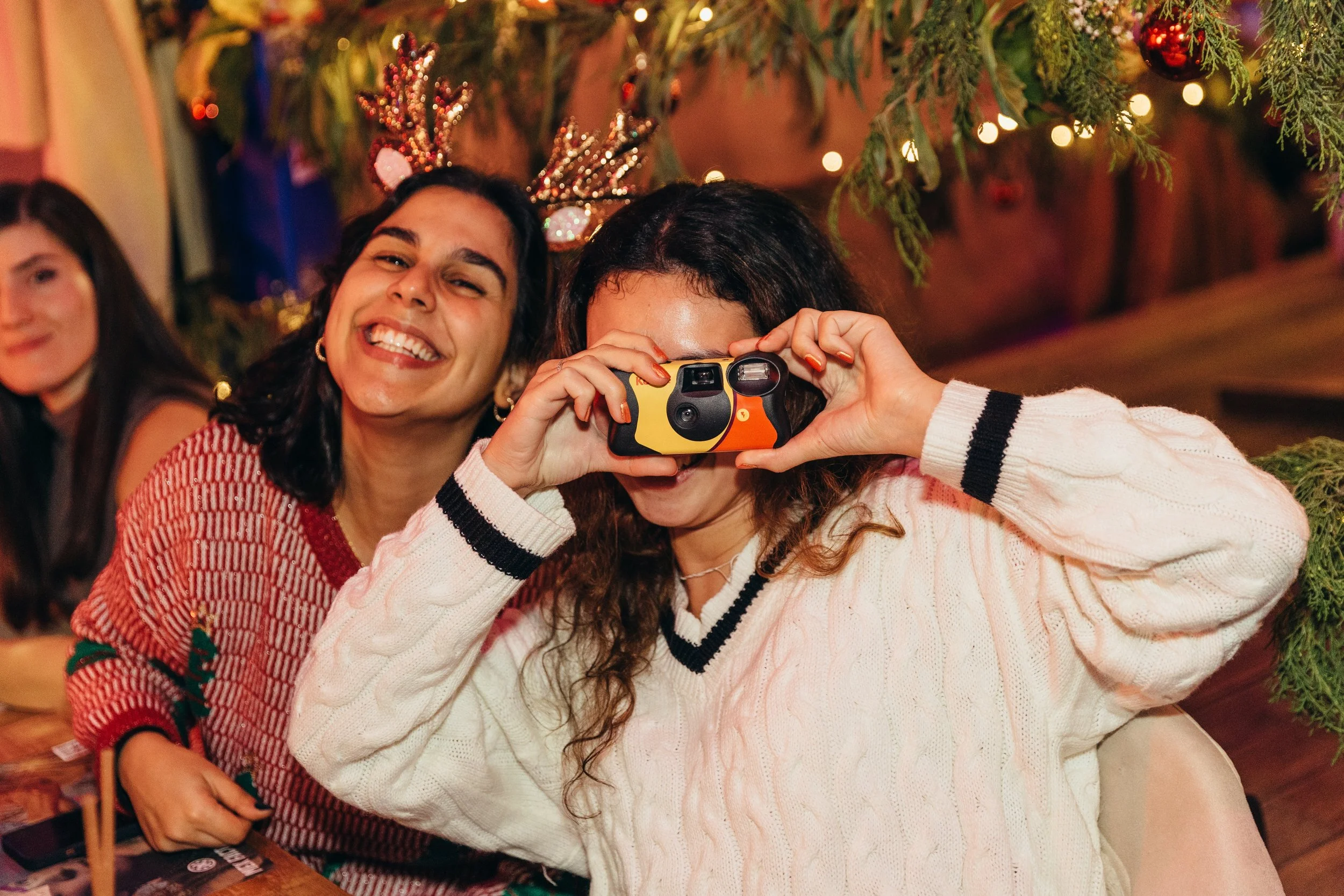Two women smiling, one holding a camera, in a festive setting with holiday decorations.