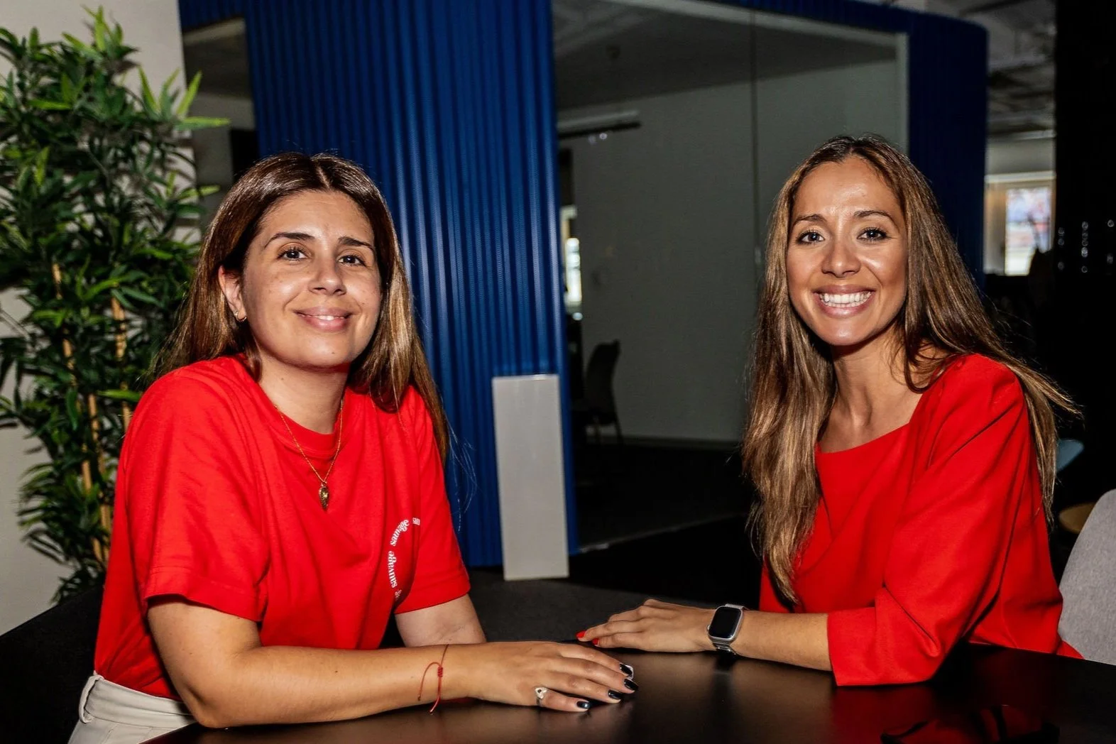 Two women sitting at a table, both wearing red tops, smiling, in an indoor setting with a plant in the background.