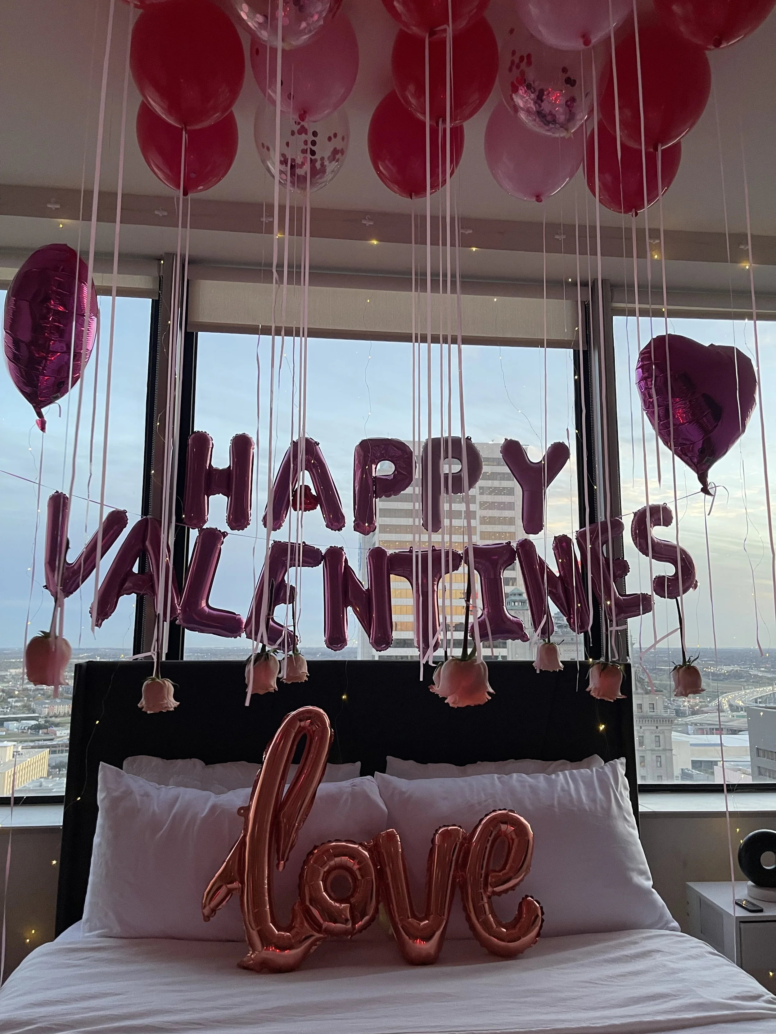 A bedroom decorated with pink and purple balloons, including thin and heart-shaped balloons, with a large 'Love' balloon on the bed. Above the bed, pink foil balloons spell out 'HAPPY VALENTINES,' and there are hanging pink roses and string lights. The window shows a cityscape with tall buildings.