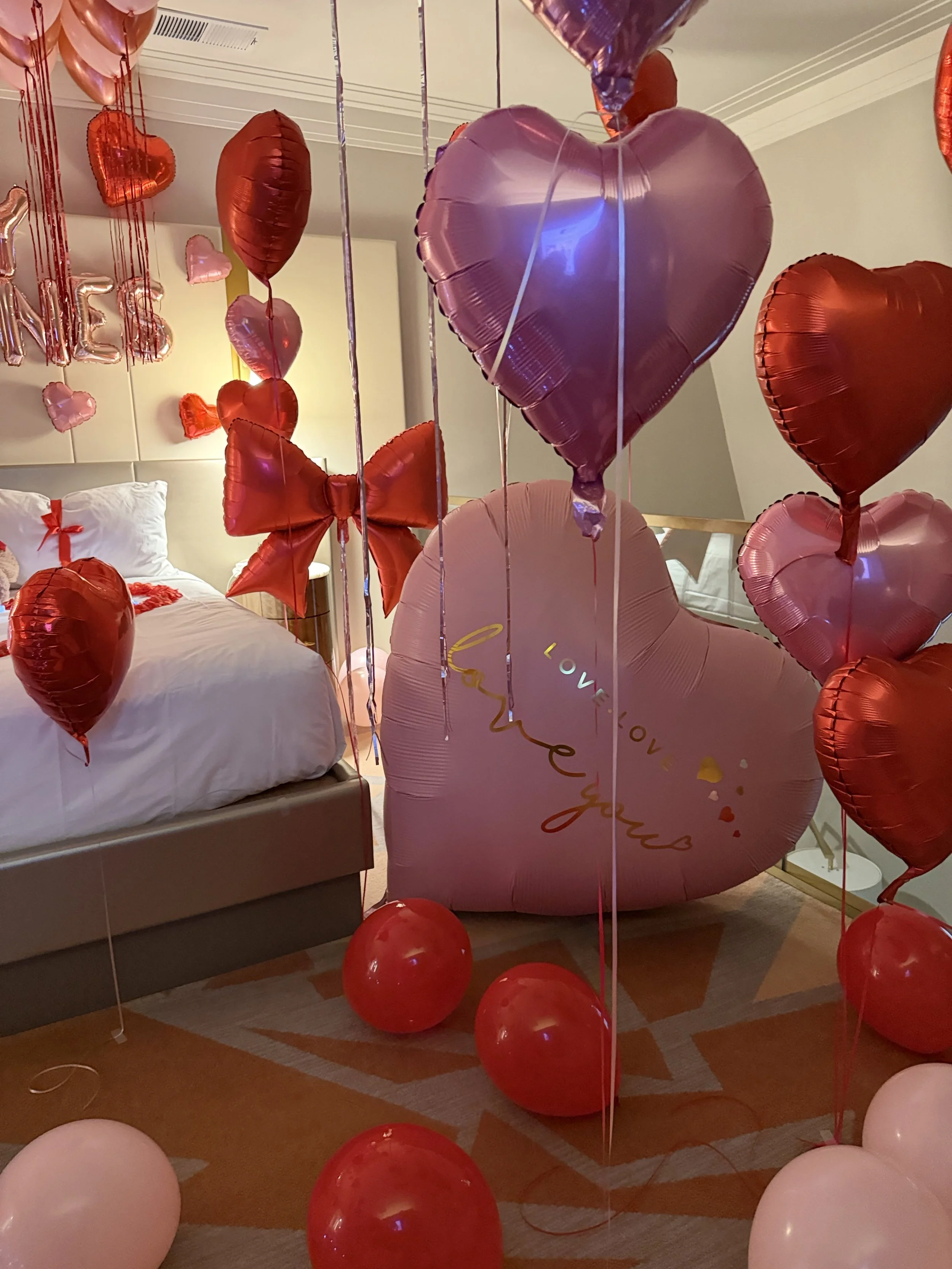 pink and red balloons decorating a hotel room. with a giant red bow balloon
