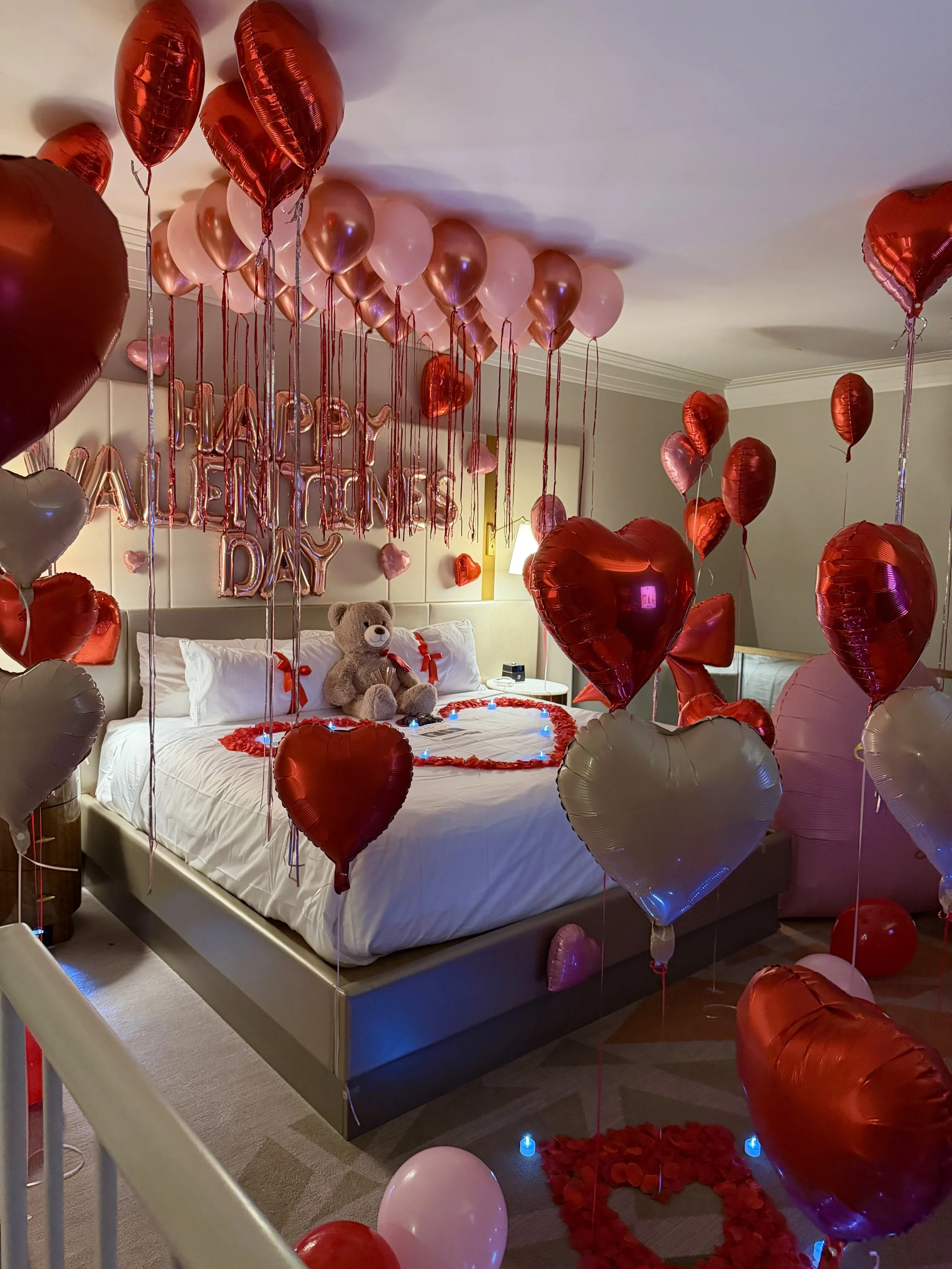 Fully decortated hotel roo with dim lighted candles. heart balloons all over and a Happy Valentines Banner with rose petal designs on the bed shaped into a heart