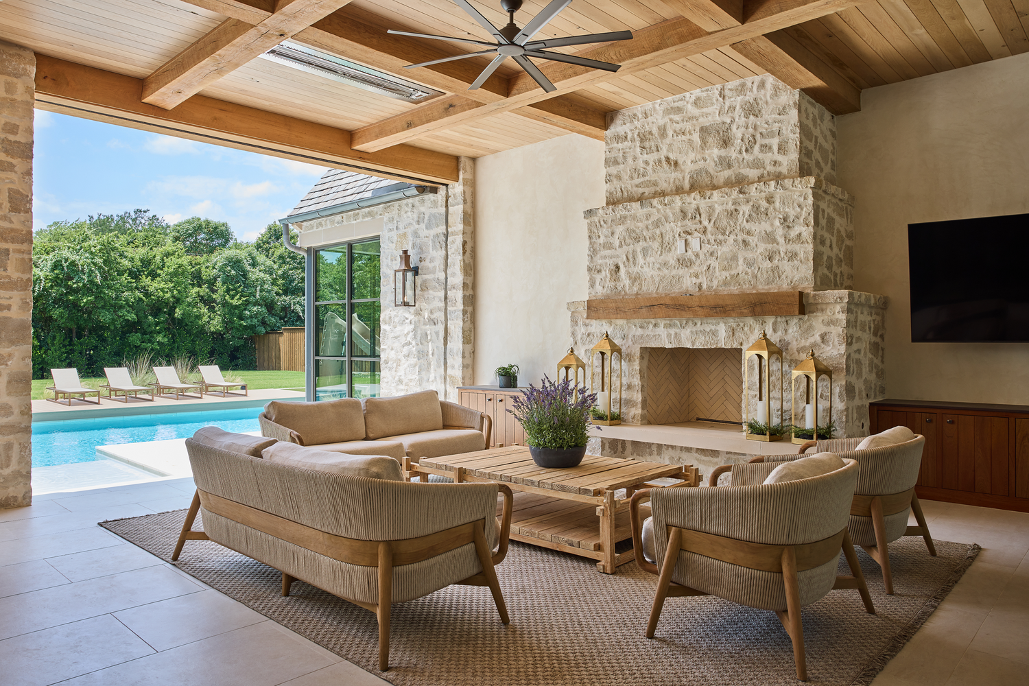 Luxury outdoor living design with covered patio seating, stone fireplace, wood ceiling beams, and poolside entertaining space.