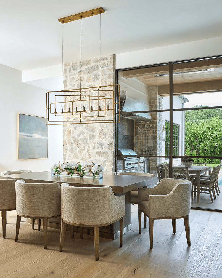 Dining room with upholstered chairs, statement chandelier, stone fireplace wall, and sliding doors opening to an outdoor entertaining area.