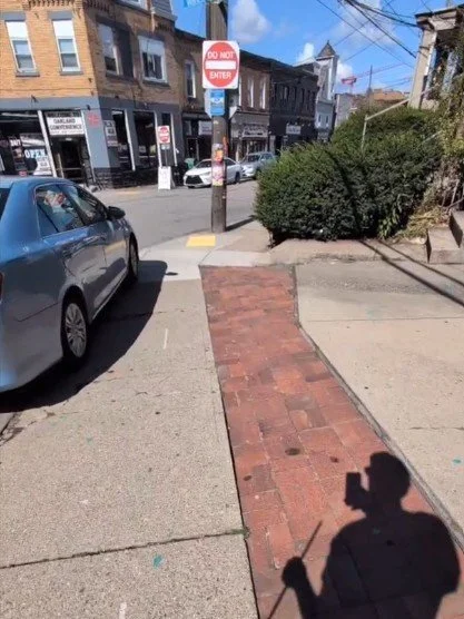 View of sidewalk with a car parked on it, and a shadow of the person taking the photo with their phone