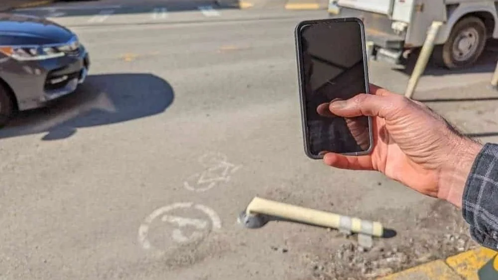 An image of someone holding a smartphone to take a photo of a broken flex post on a pittsburgh street.
