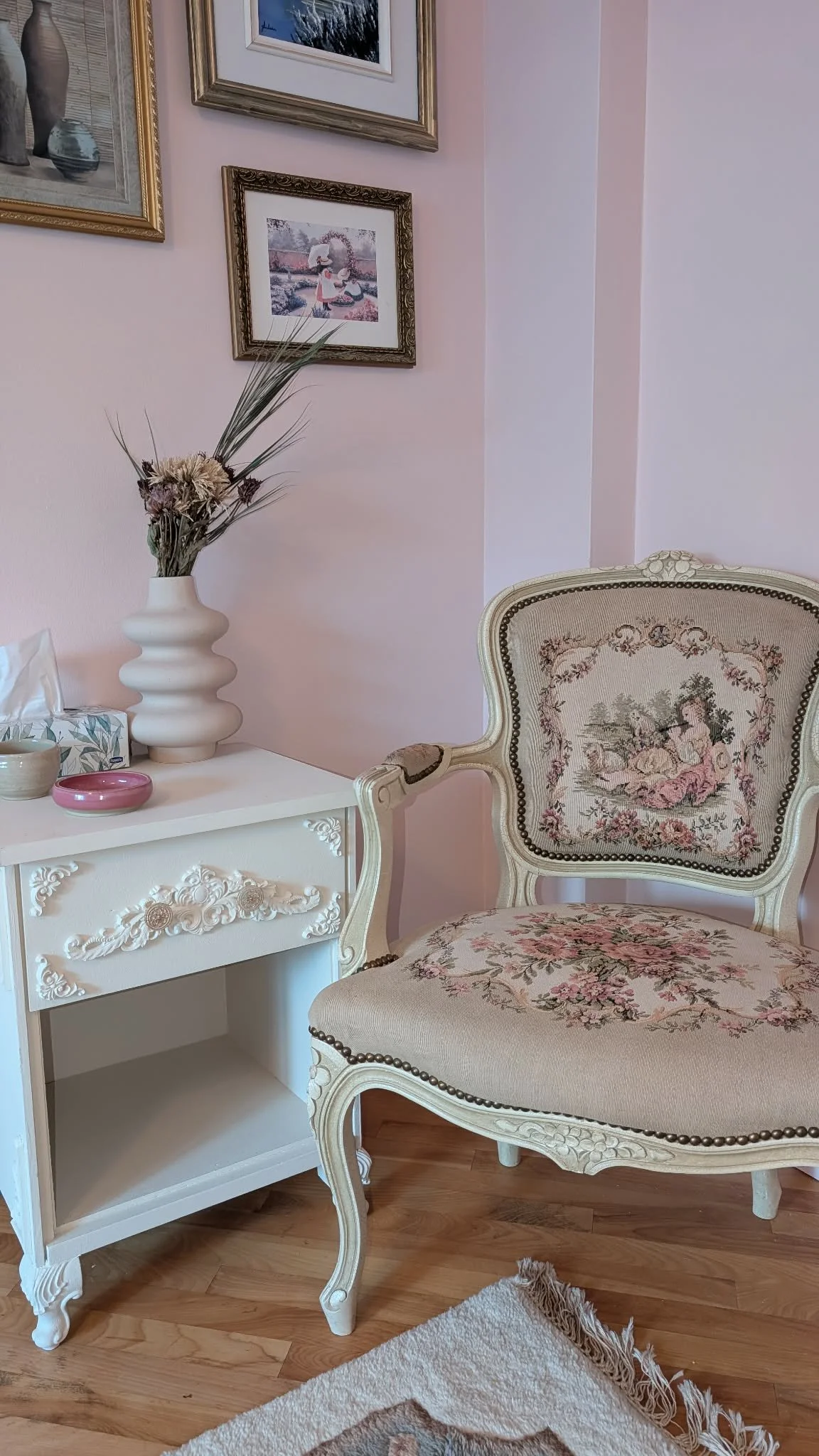 Un coin de chambre avec un fauteuil à motif floral, une petite table blanche avec des décorations, un vase avec des fleurs séchées, plusieurs tableaux décoratifs sur le mur rose et un tapis blanc et beige sur le sol en bois.
