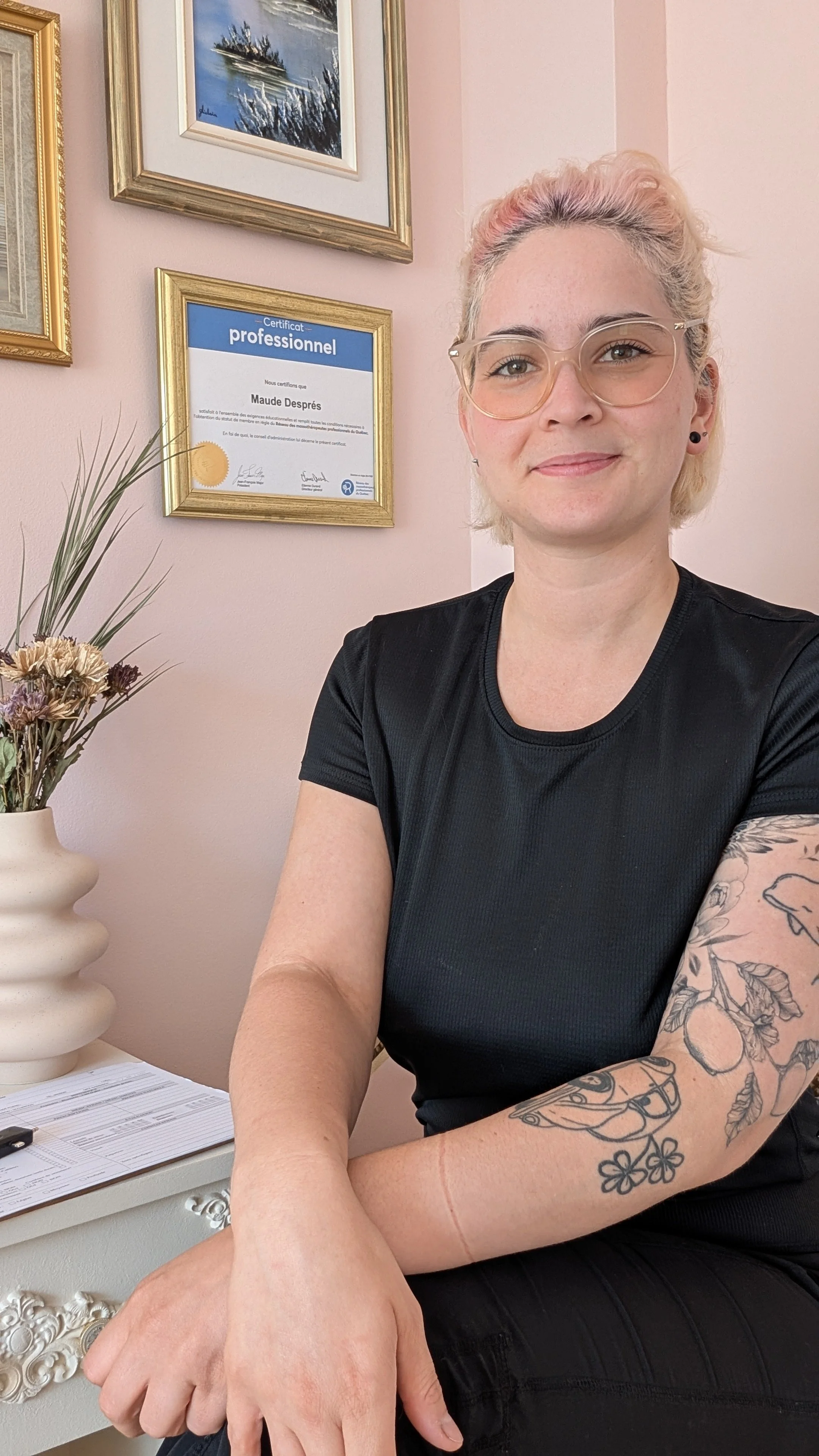 Une femme souriante portant des lunettes, avec des tatouages sur le bras, des cheveux teints rose, assise à côté d'une table de chevet beige avec un bouquet de fleurs séchés et deux certificats encadrés sont accrochés au mur derrière elle.