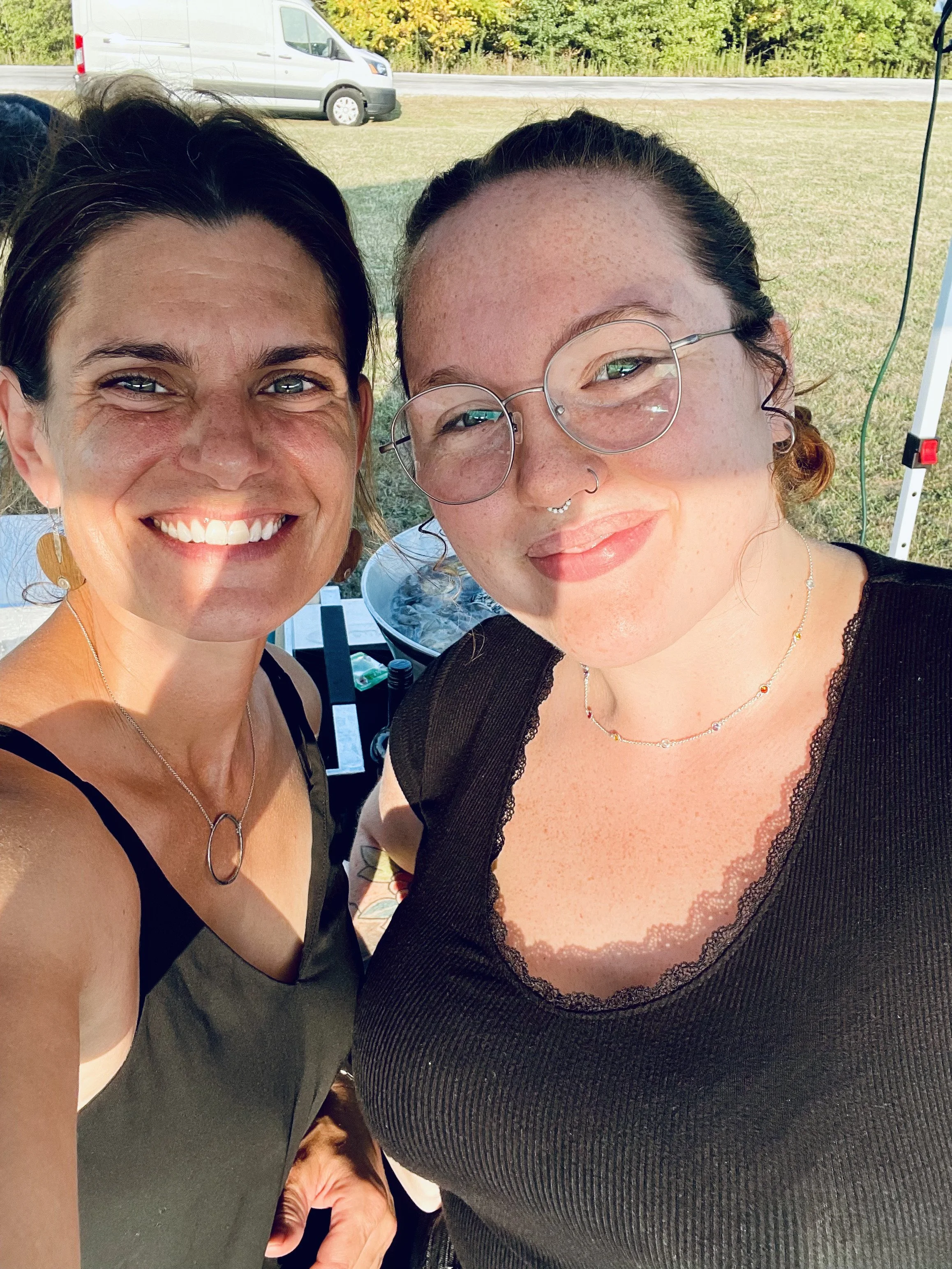 Two women taking a selfie outdoors, smiling. One has short dark hair and is wearing a sleeveless top and earrings. The other has glasses, freckles, and is wearing a black top with lace trim.