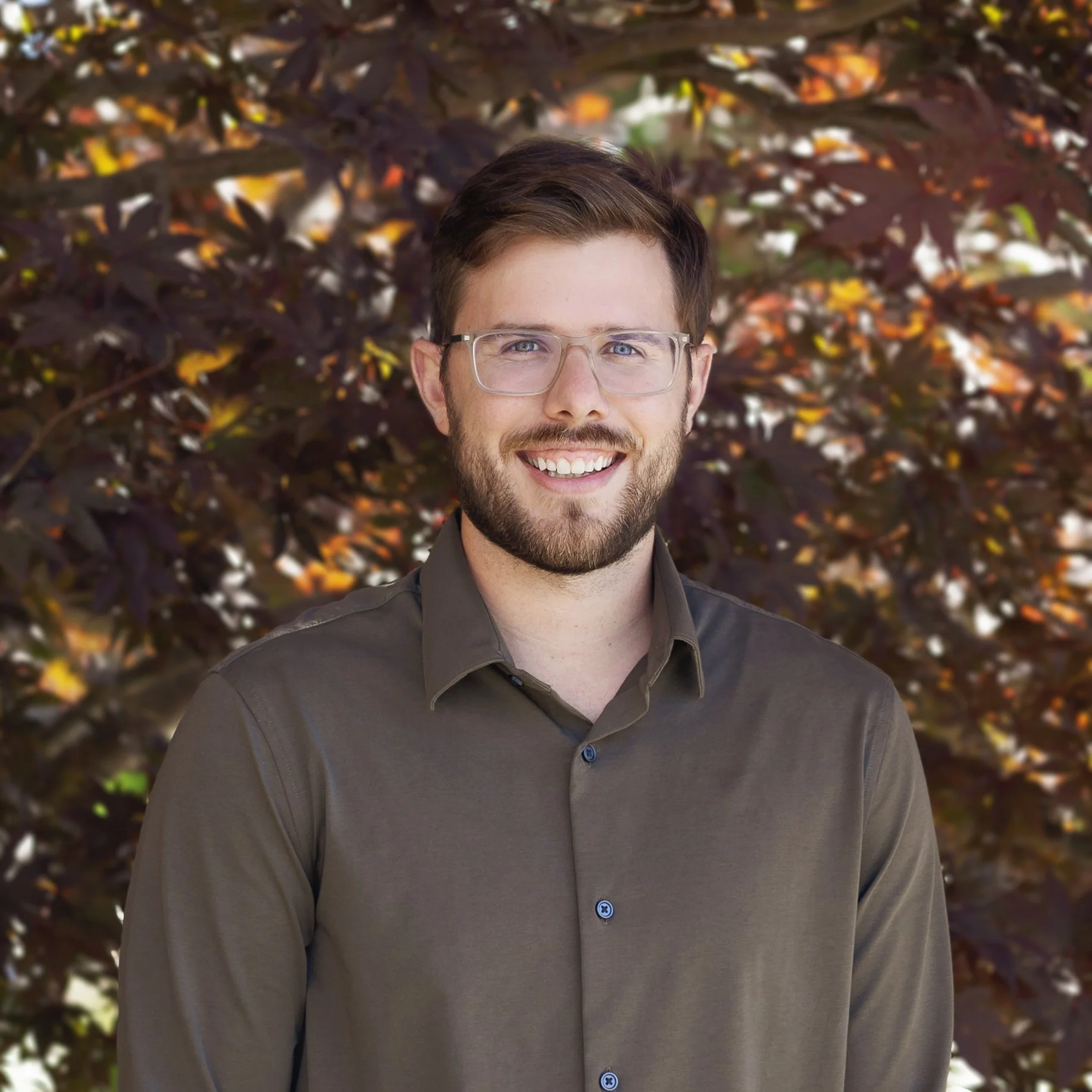 A man with glasses and a beard is smiling, wearing a green shirt, and standing in front of a bush with red and orange leaves.