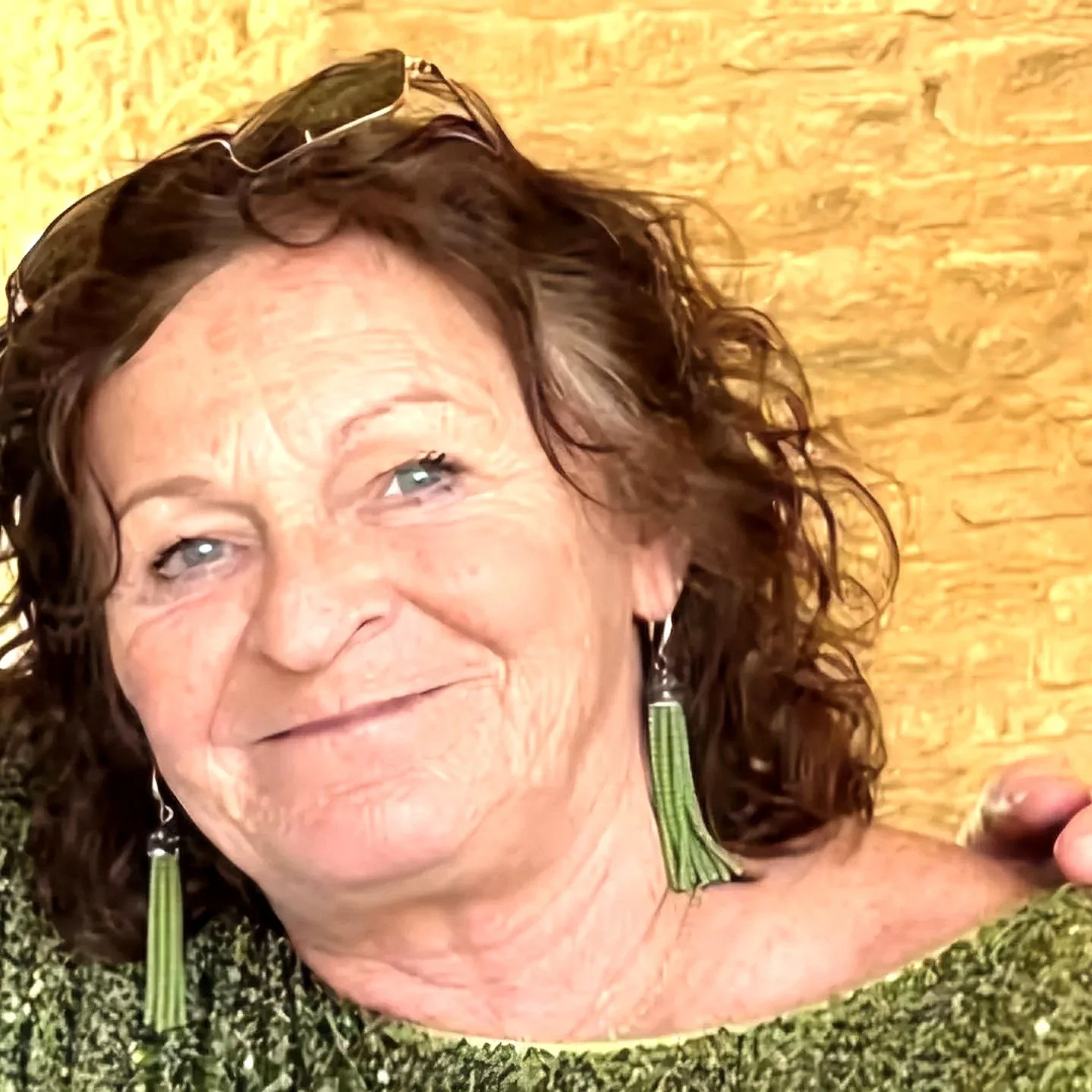 Close-up of an elderly woman with curly brown hair, wearing green earrings, and a green knitted top, smiling in front of a wooden background.