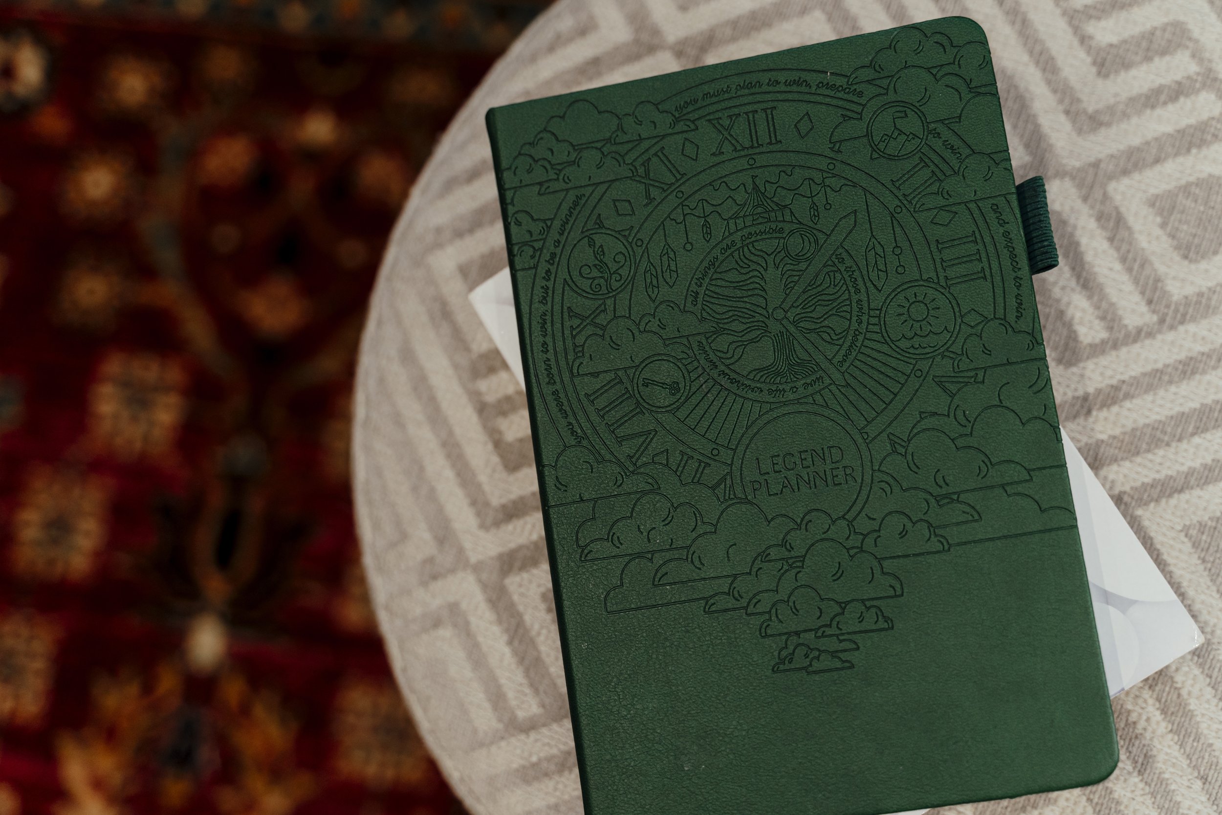 A green planner titled 'Legend Planner' with intricate circular and cloud designs on the cover, resting on a textured beige fabric surface.
