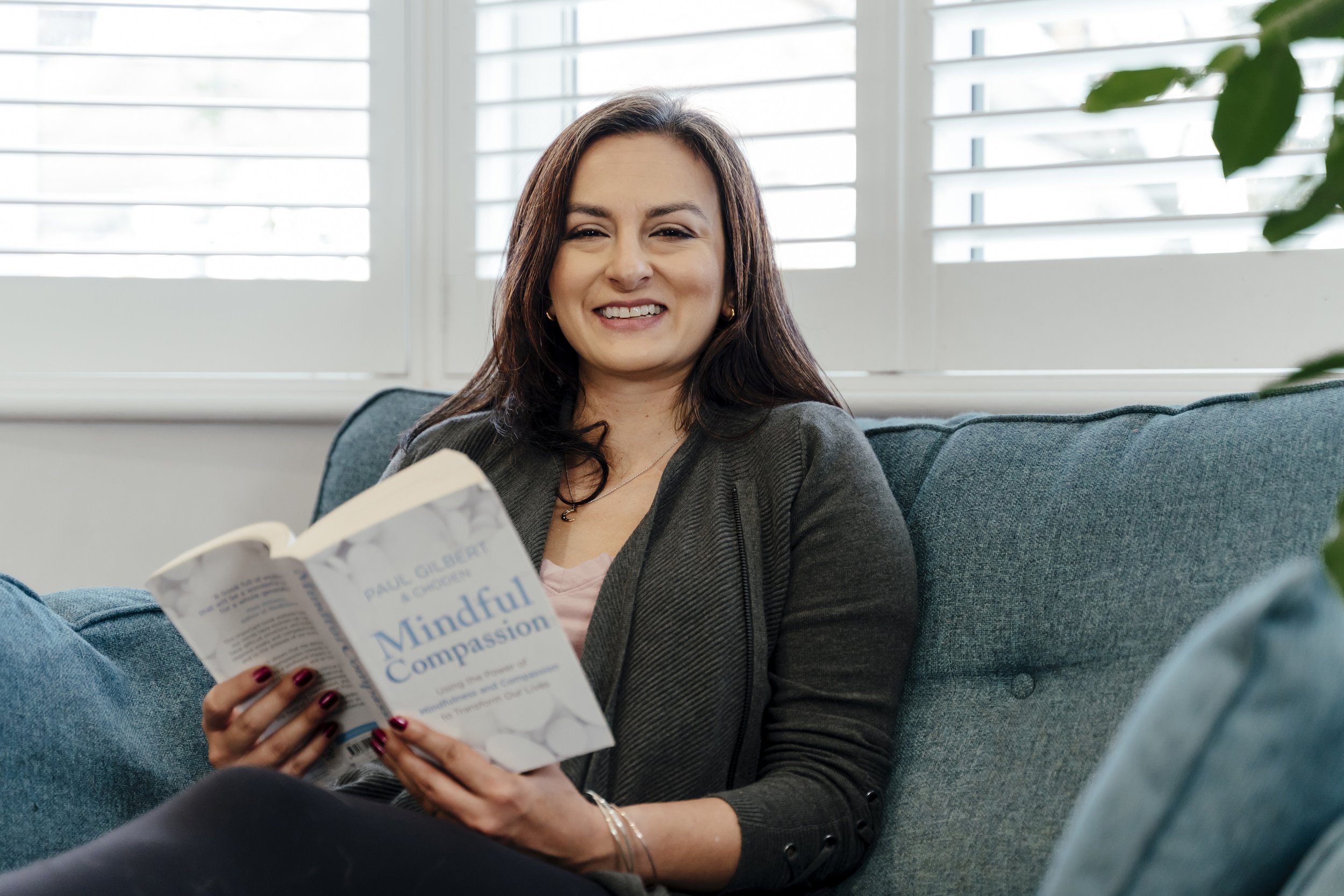 Smiling woman sitting on a blue couch, holding a book titled "Mindful Compassion" by Paul Gilbert, in a bright room with white window blinds.