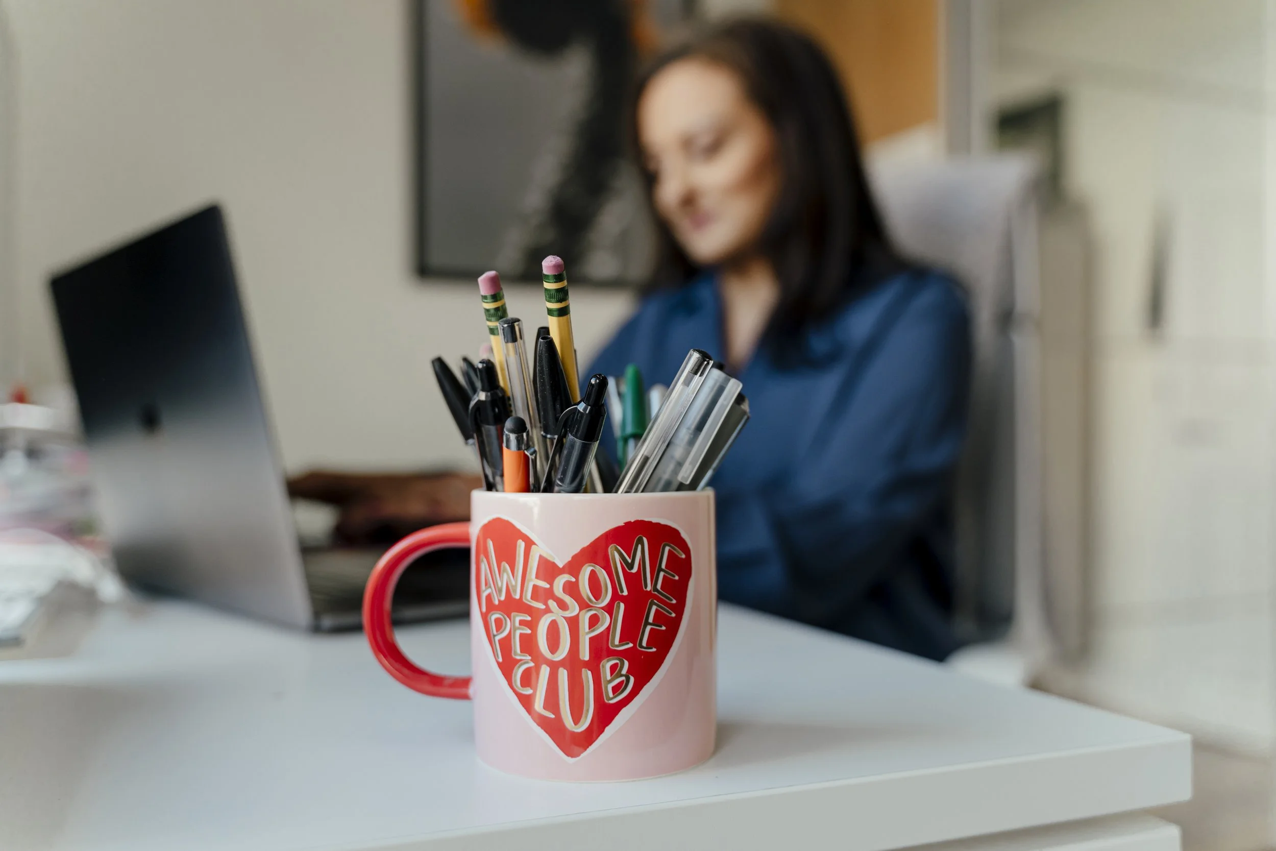 A pink mug with a red heart and the words 'Awesome People Club' on it, filled with pens and pencils, on a white desk in an office setting, with a woman working in the background.