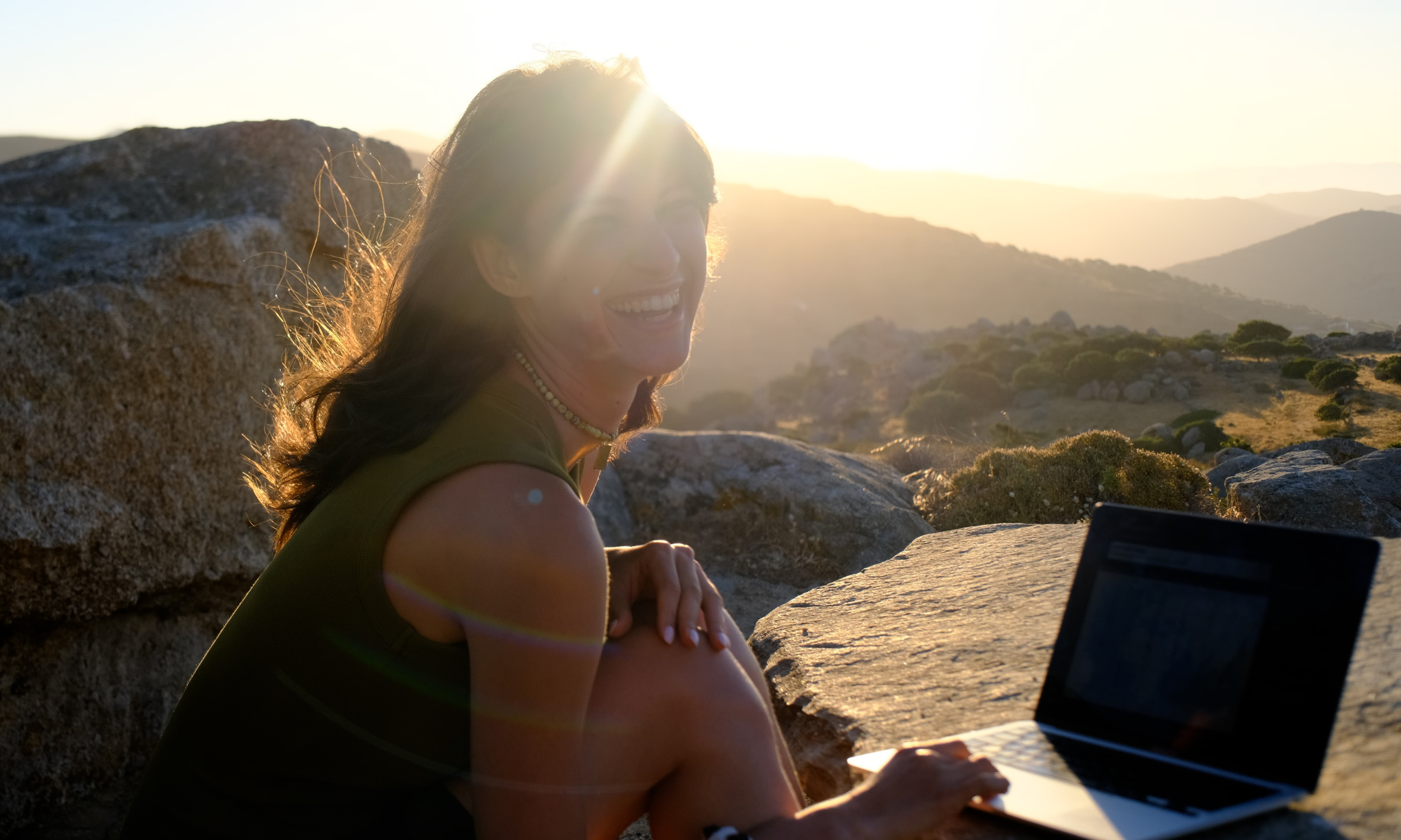 A woman with brown hair smiling while sitting on rocks outdoors during sunset, working on a laptop.
