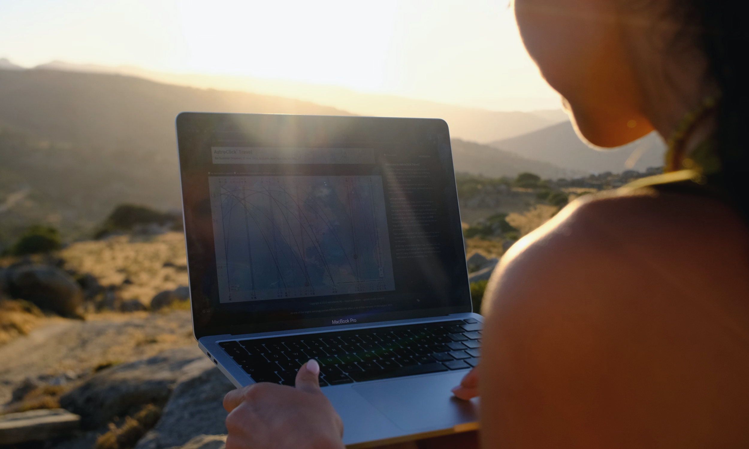 A person is sitting outdoors on rocks at sunset, using a MacBook Pro with a screen displaying a graph or chart. The person is partially visible, focusing on their shoulder and hand.