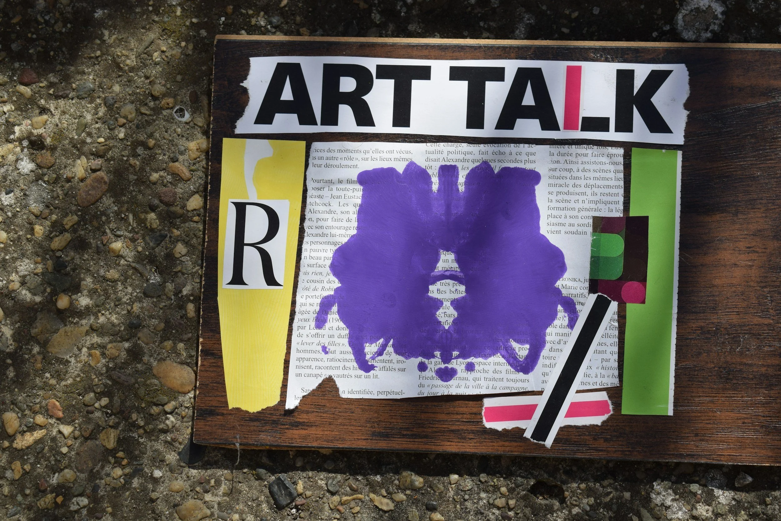 A piece of black wooden board with various stickers and paper attached to it, including a large purple paint splash, the word 'ARTTALK' in black and red letters, colorful geometric strips, and text snippets, placed on a gravel surface.