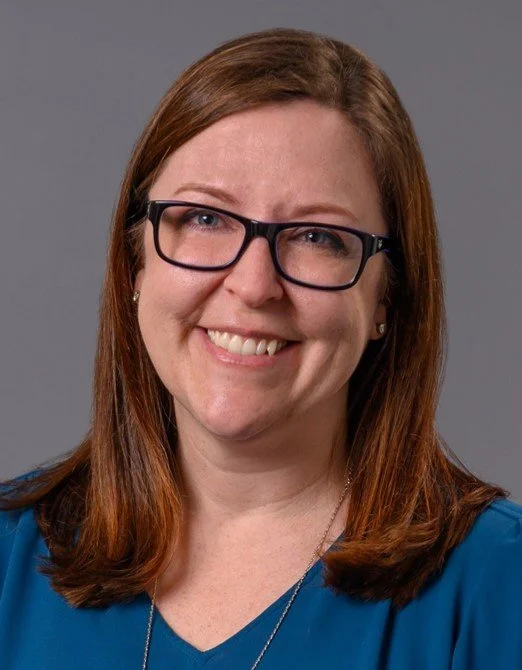 A woman with long red hair wearing glasses and a blue blouse, smiling at the camera against a gray background.