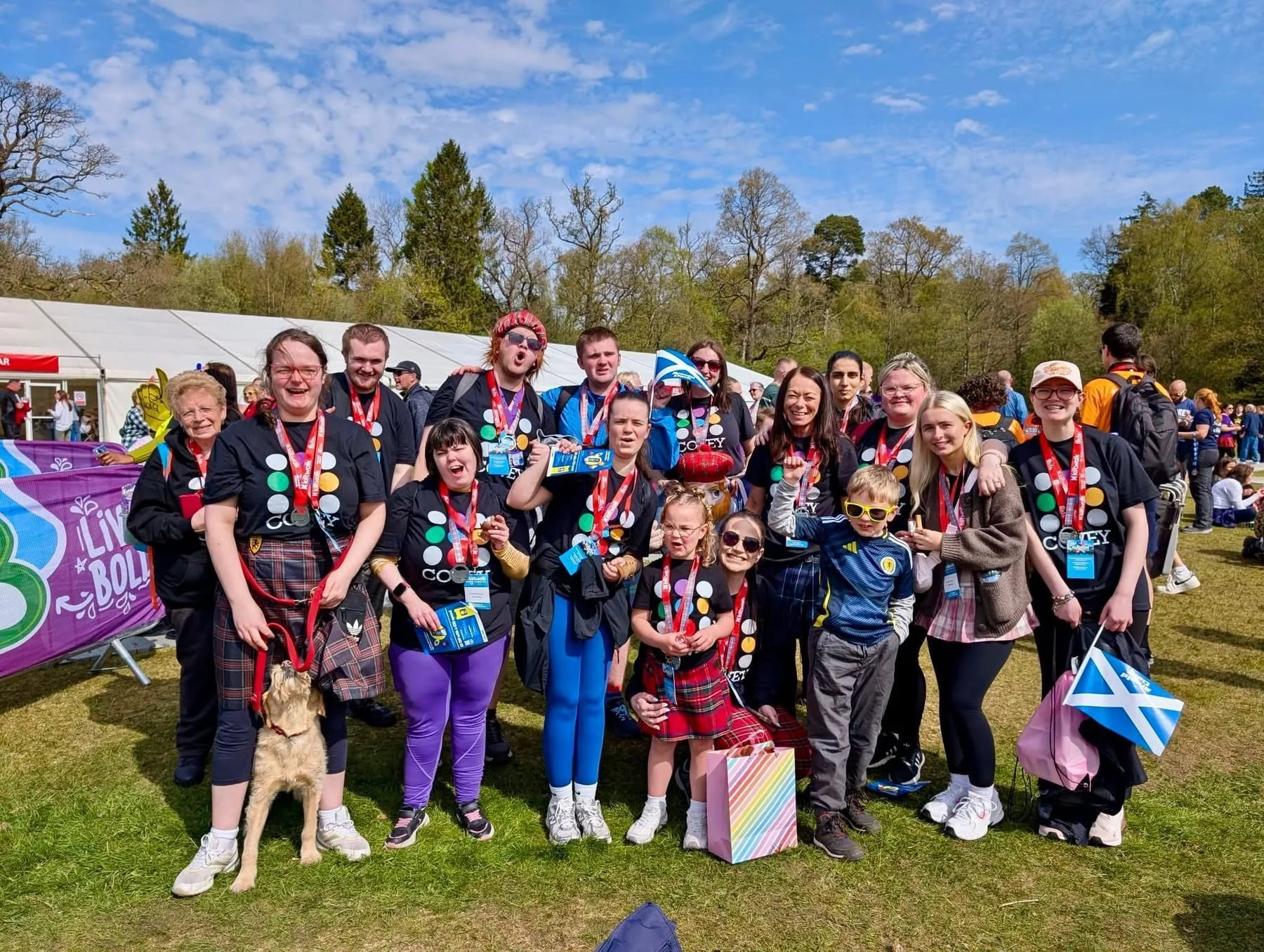 A large group of people outdoors at a festival or event, many wearing black shirts with colorful dots and lanyards, some holding flags or banners, and a dog on a leash in the front.