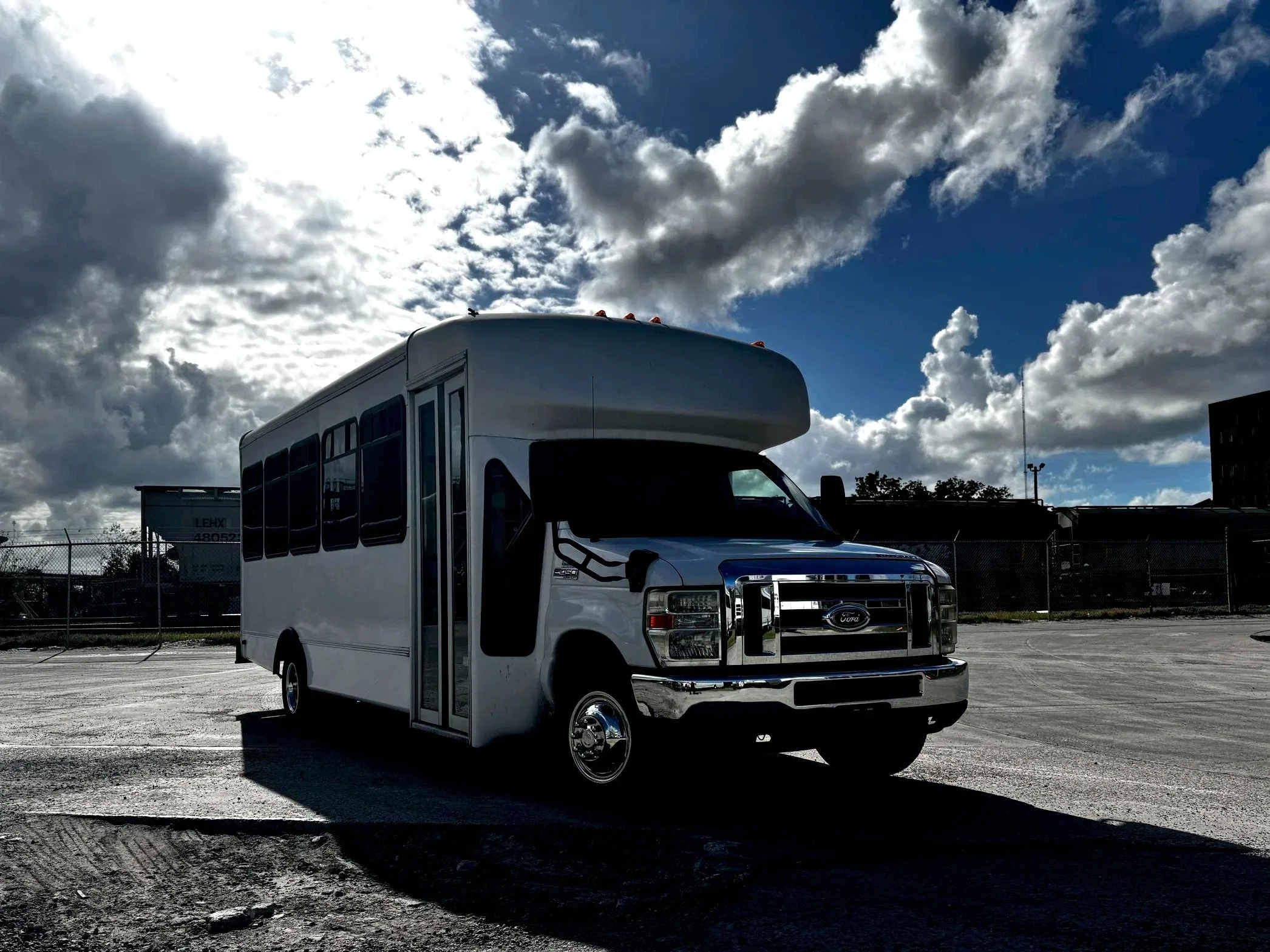 A small white limo bus parked on an open lot with a chain-link fence and some buildings in the background, under a partly cloudy sky with sunlight breaking through clouds.