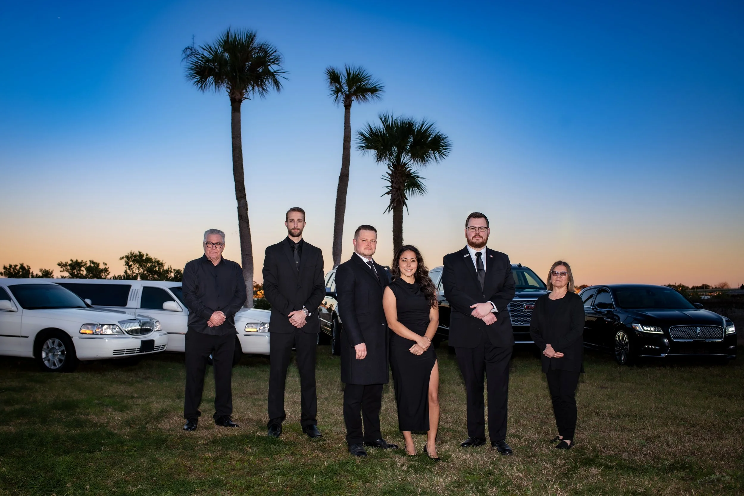 Group of six people in formal black attire standing outdoors in front of luxury cars and palm trees at sunset.