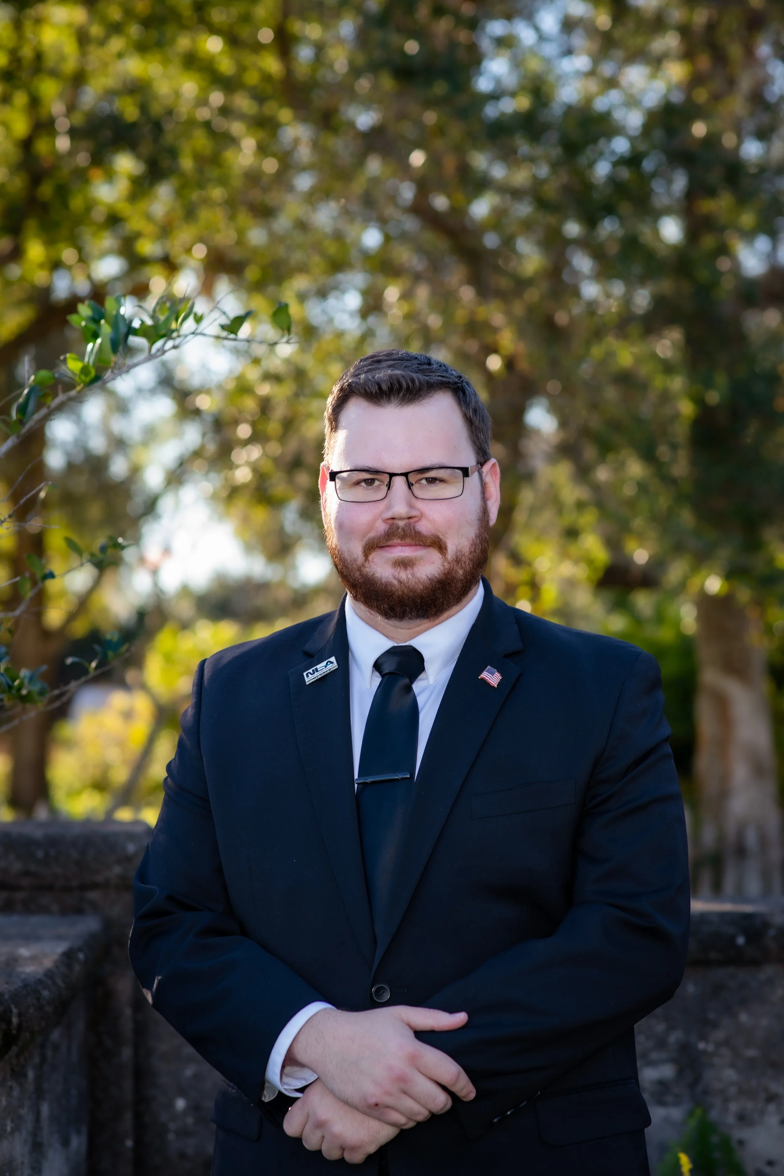 A man with glasses and a beard wearing a black suit, white shirt, and black tie, standing outdoors in front of green trees, with some pins on his lapel.