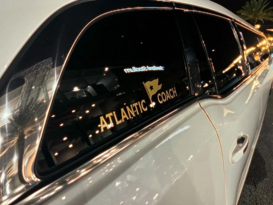 Close-up of a white car window with a decal that reads "Atlantic Coach" and features a star and flag graphic, taken at night with reflections of lights.