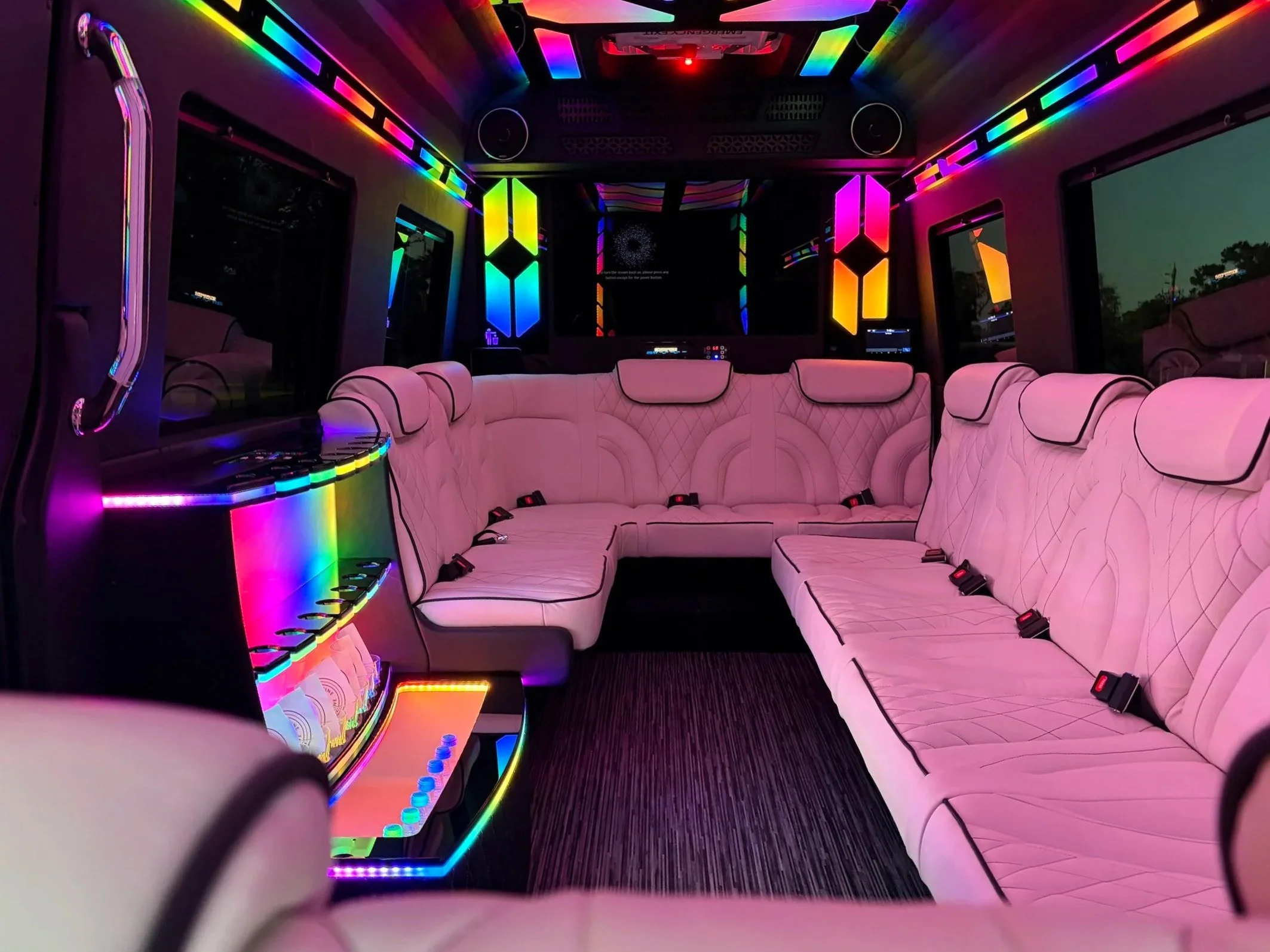 Interior of a bus or RV with pink quilted seats, colorful LED lighting, and windows showing trees outside