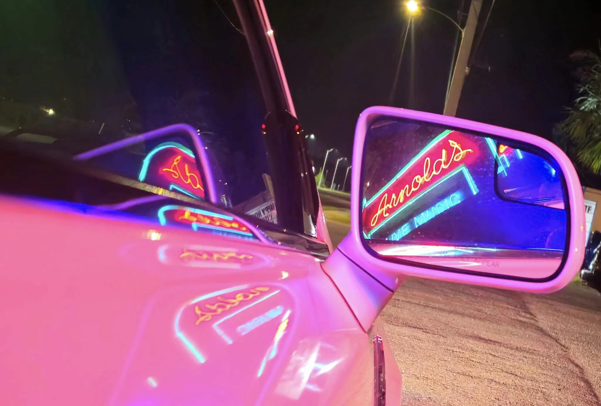 Pink car's side mirror reflecting neon sign for Arnold's live music at night.