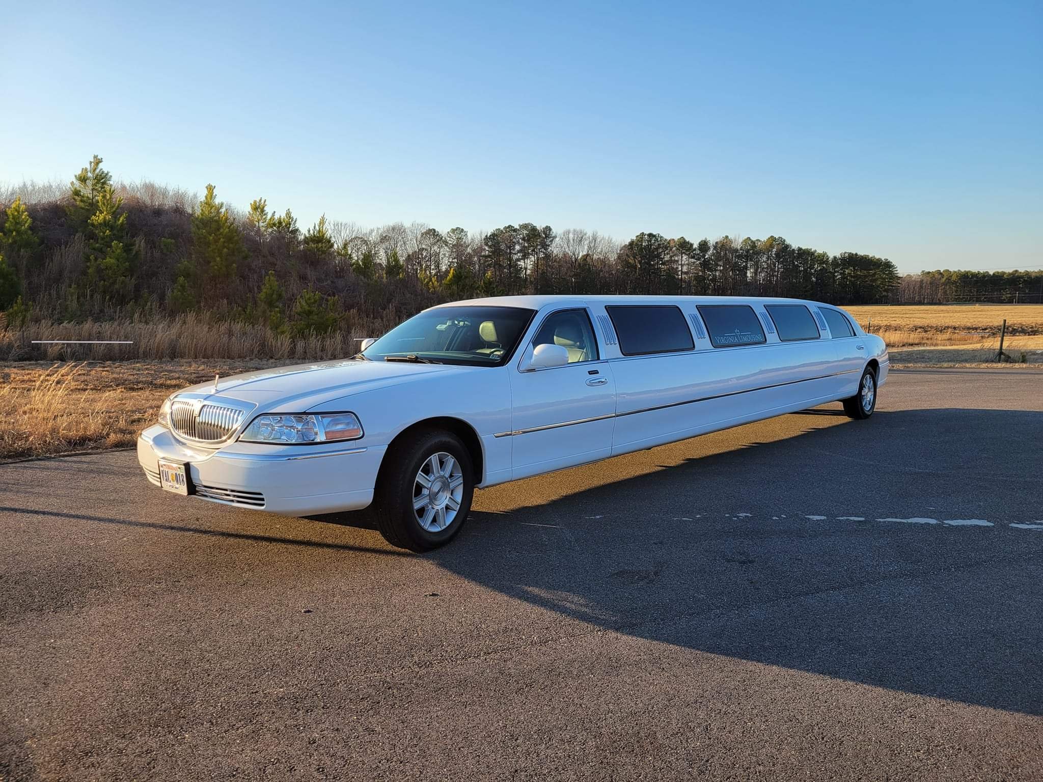 A white limousine parked on the side of a road during sunset, with trees and open fields in the background.