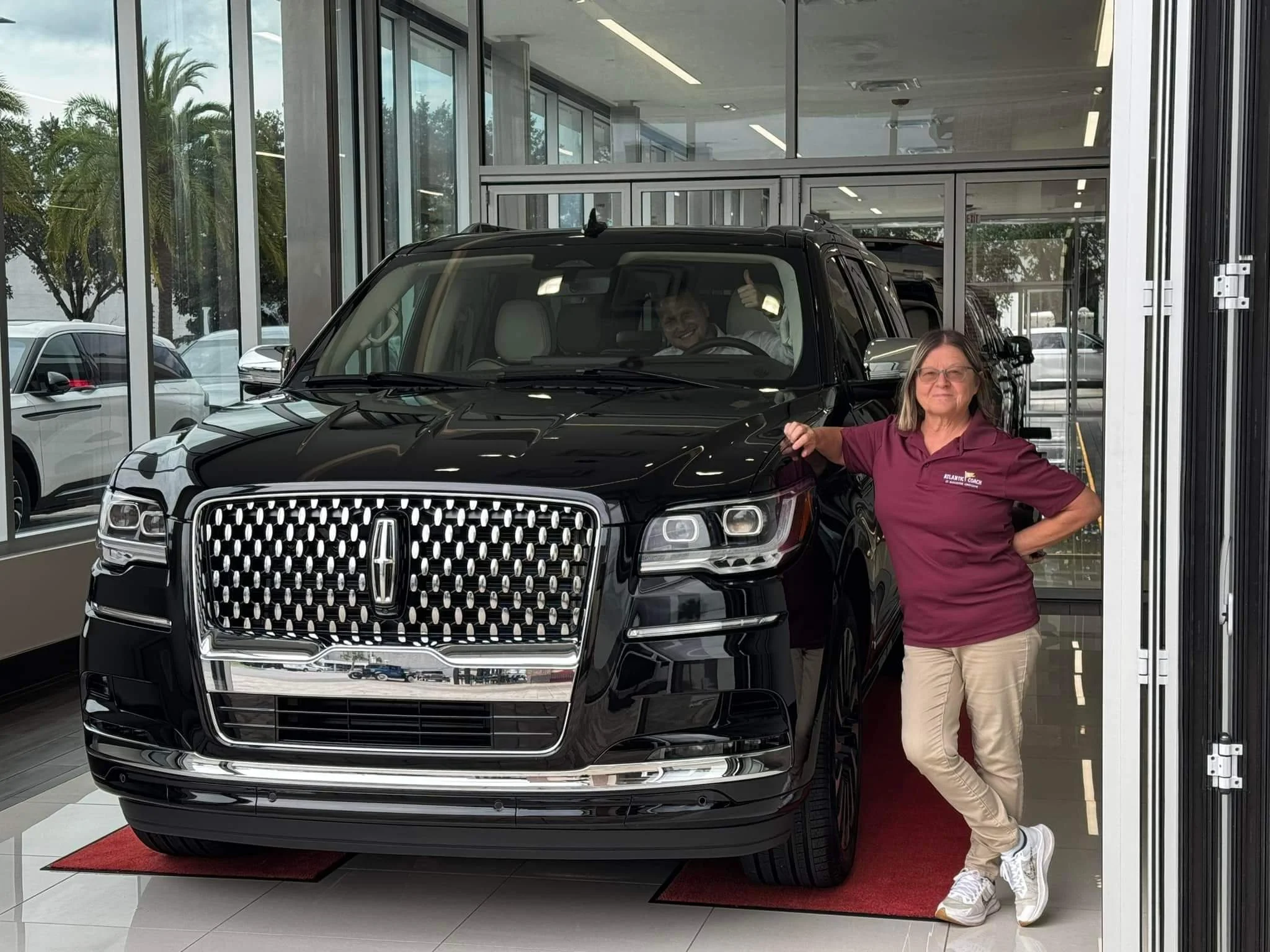 A woman in a maroon shirt and beige pants standing next to a black Lincoln SUV inside a car dealership showroom. She is smiling and leaning against the vehicle with one hand on the hood. The driver, visible through the windshield, is giving a thumbs-