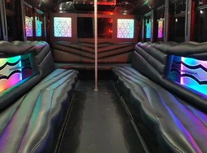 Interior of a party bus with long leather bench seats, colorful LED lights, and two screens showing static.
