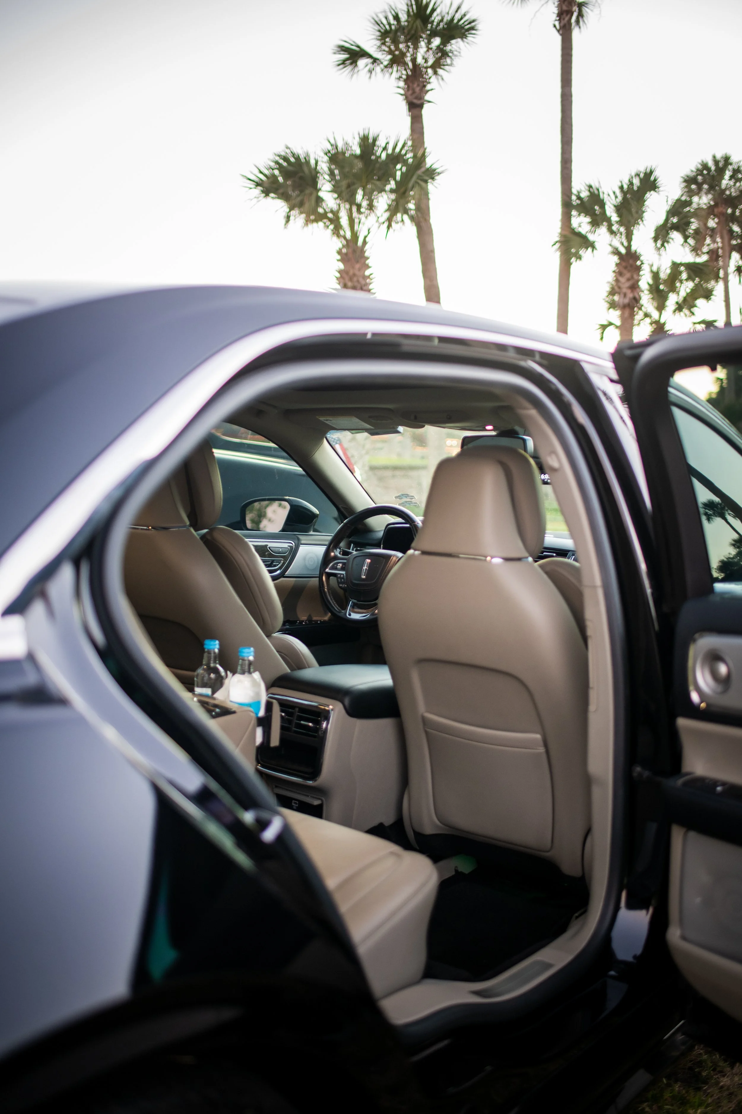 The interior of a black SUV with beige seats, water bottles in cup holders, and palm trees outside at sunset.