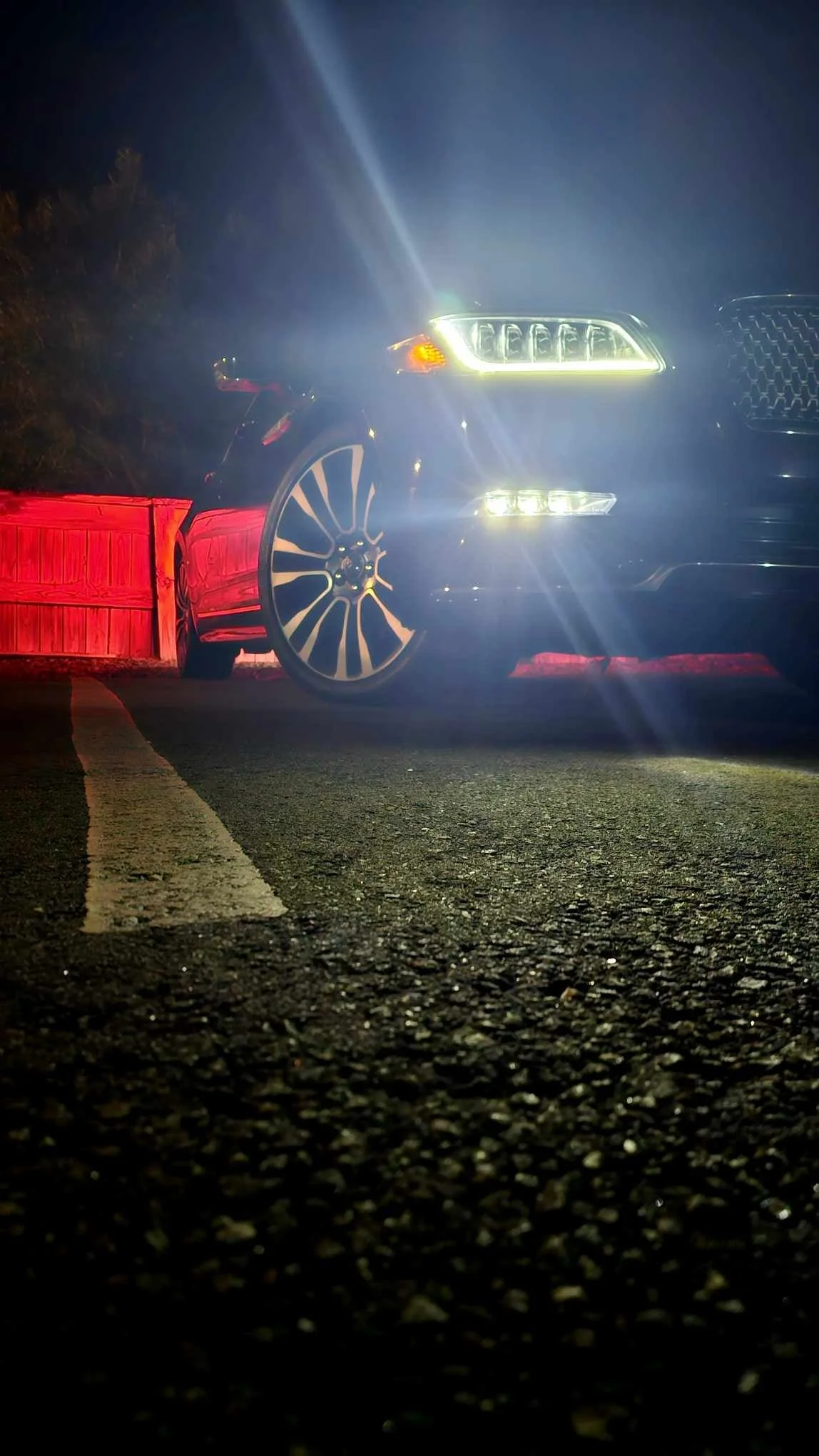 A black SUV with bright headlights parked next to a red fence at night on a paved road.