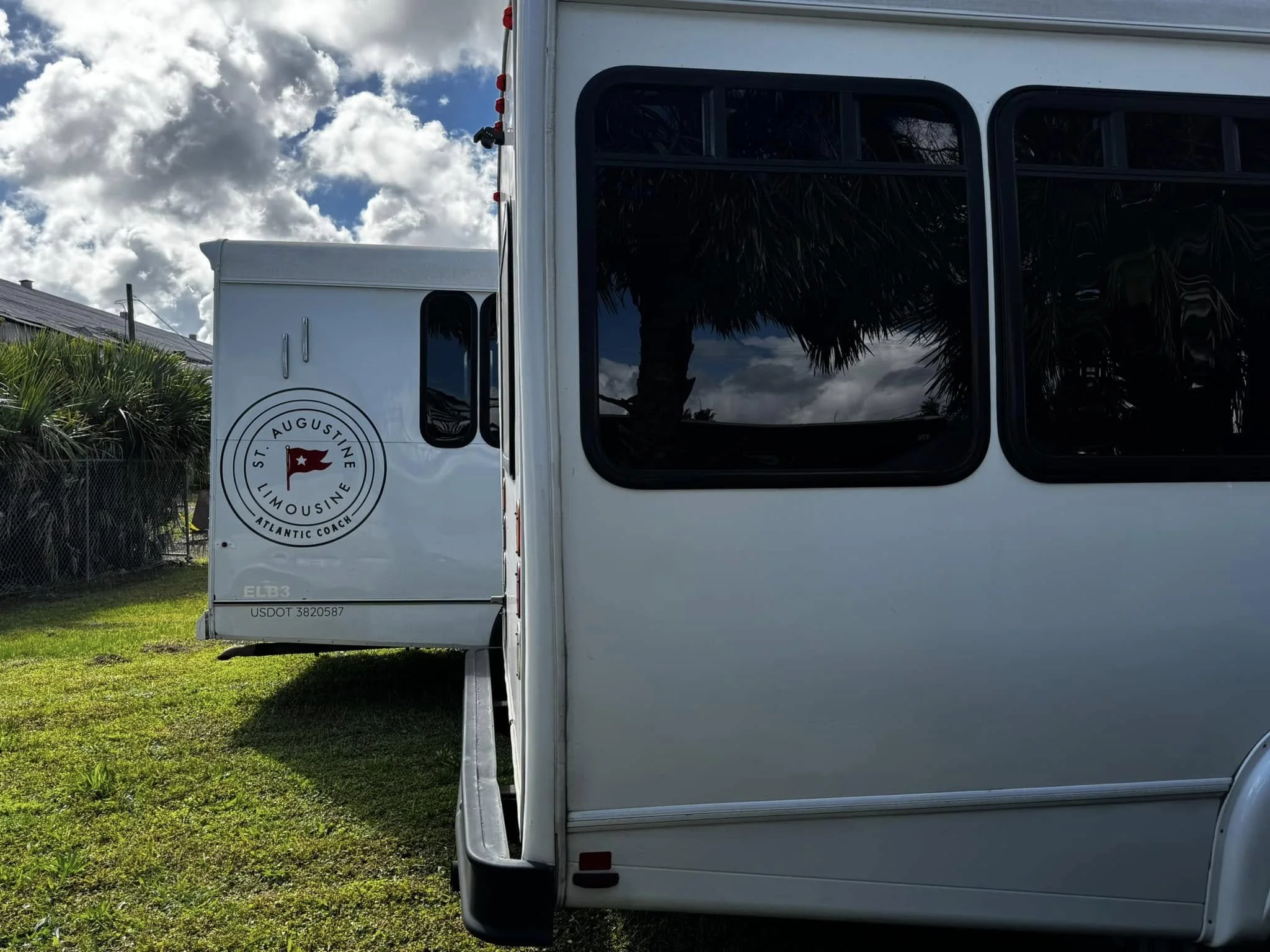 Rear view of two white RVs parked on grass, with a fence and palm trees in the background, under a partly cloudy sky.